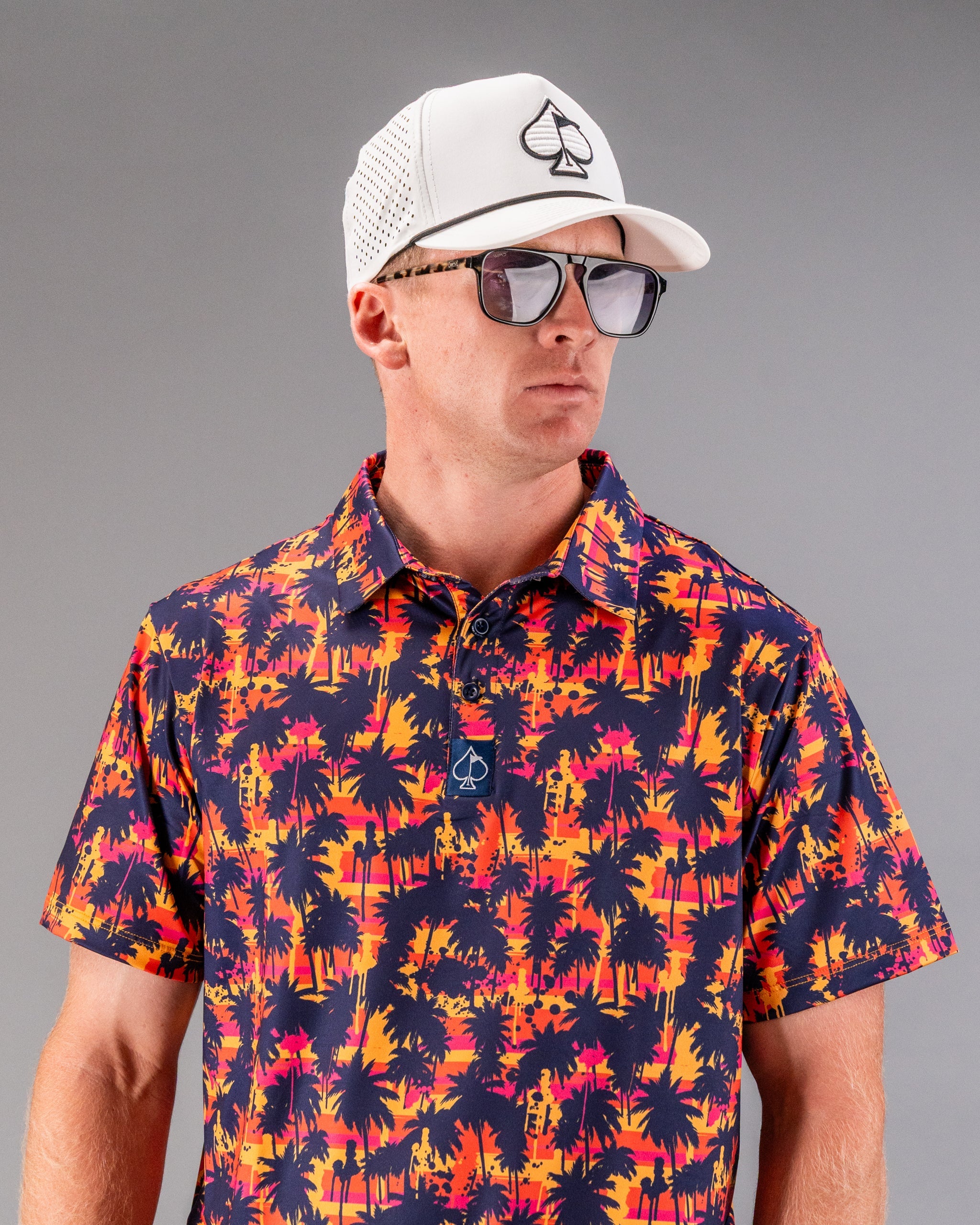Man wearing a white golf cap with a spade logo, black sunglasses, and a vibrant tropical palm tree patterned shirt in suns...