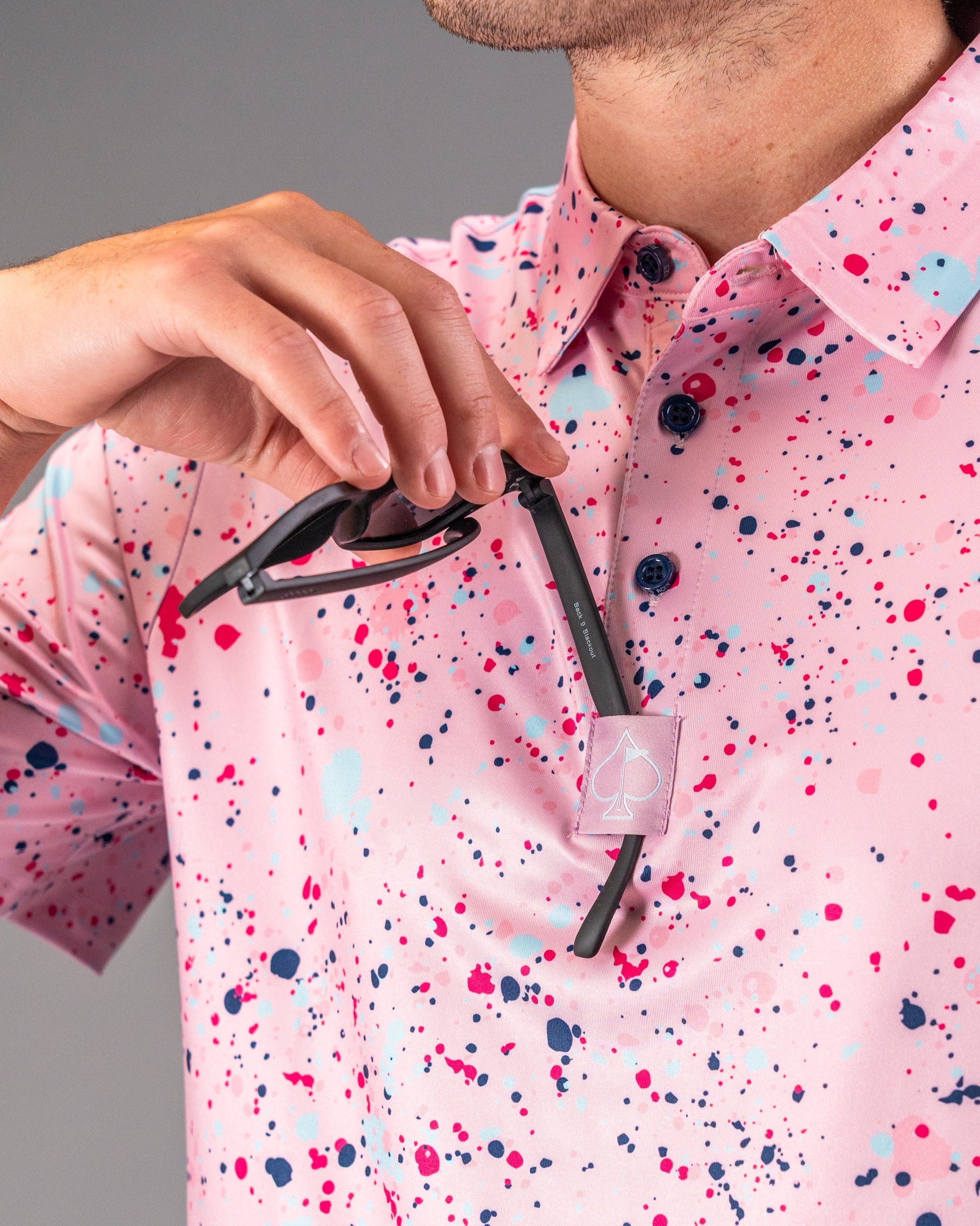Bold pink splatter golf polo shirt with colorful accents, featuring a stylish collar and sunglasses held in hand.