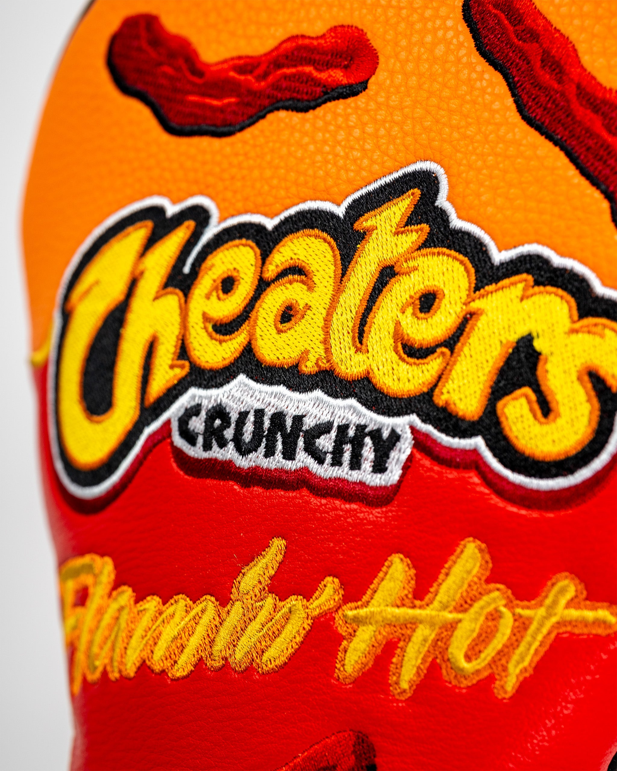 Close-up of a colorful golf driver cover featuring embroidered "Cheaters" logo, "Crunchy" and "Chili Hot" text with chili pepper graphics.