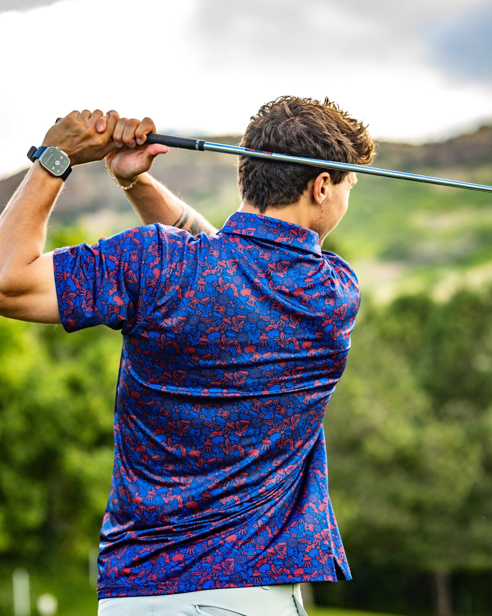 Man swinging a golf club, wearing a navy shirt with a colorful elephant pattern, set against a scenic outdoor background.