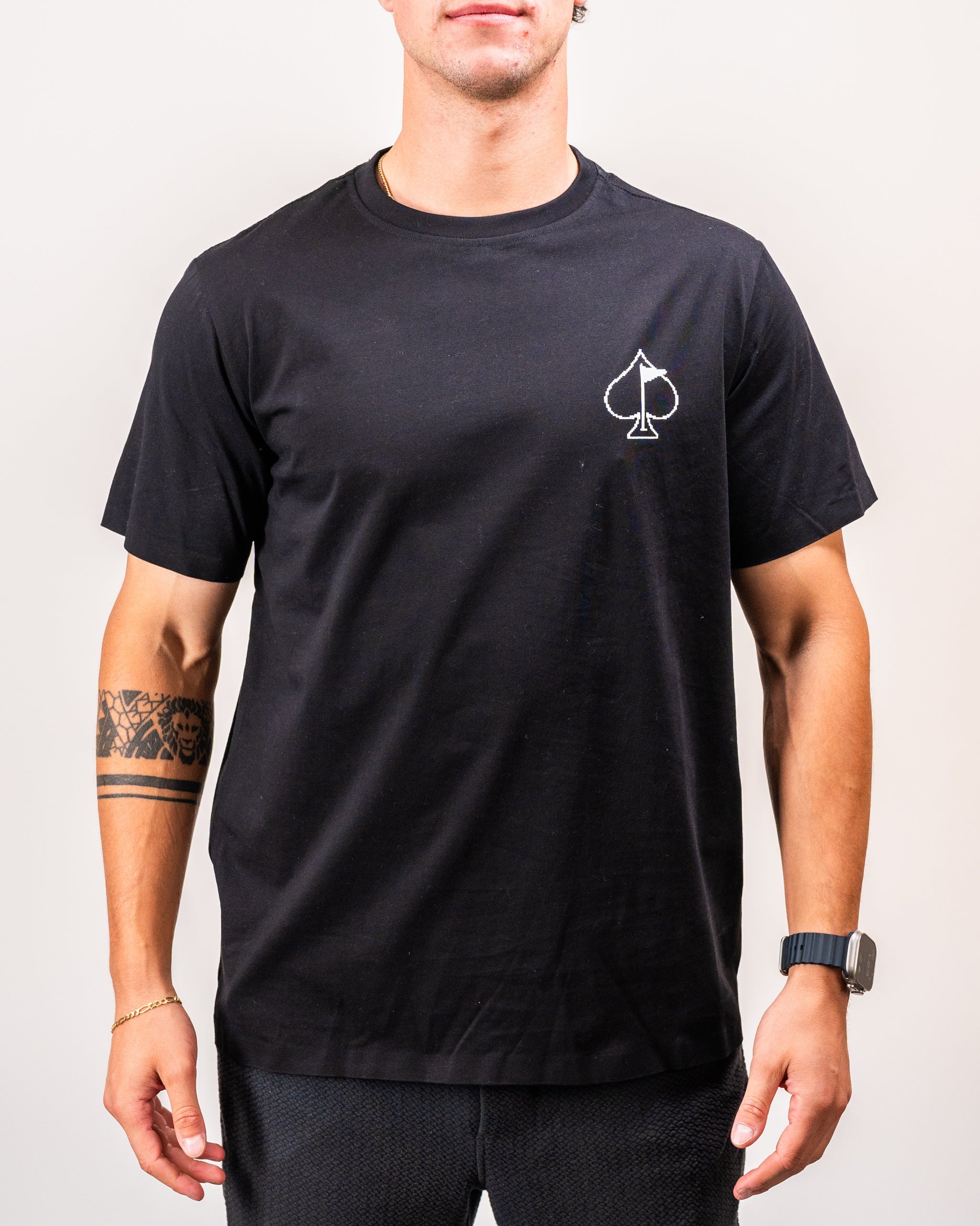 Man wearing a black T-shirt featuring a small white 8-bit spade symbol on the chest, with visible tattoos on his left arm and wearing a black watch.