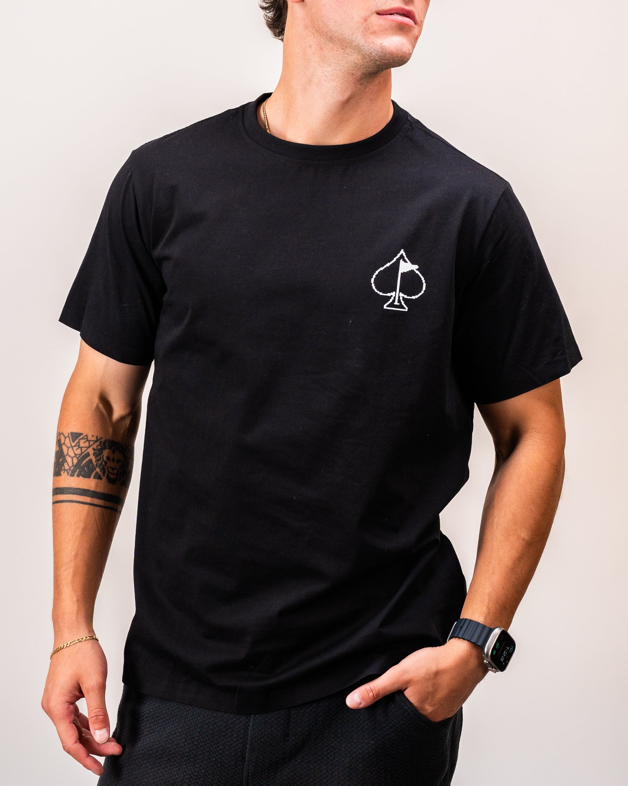 Man wearing a black t-shirt featuring a white spade symbol on the chest, styled with a casual wristwatch and displaying a tattoo on his left arm.
