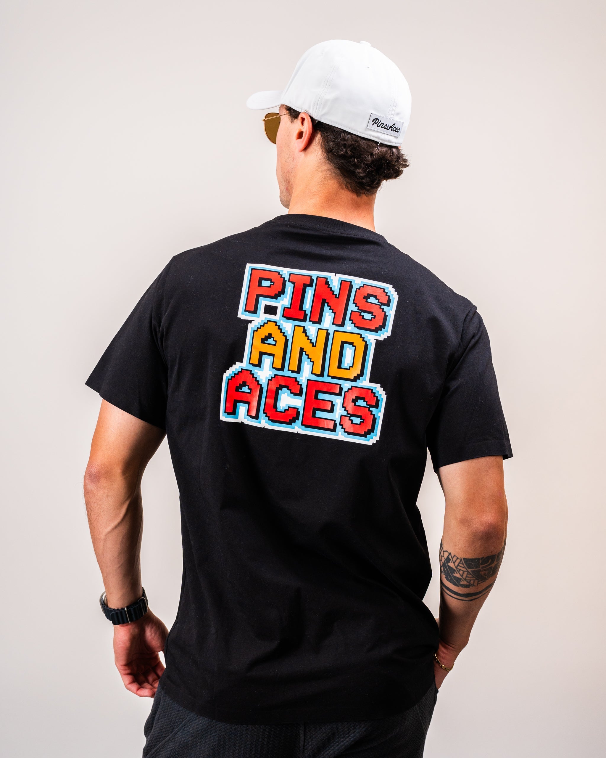 Man wearing a black t-shirt with "PINS AND ACES" in colorful 8-bit lettering on the back, and a white cap facing away from the camera.