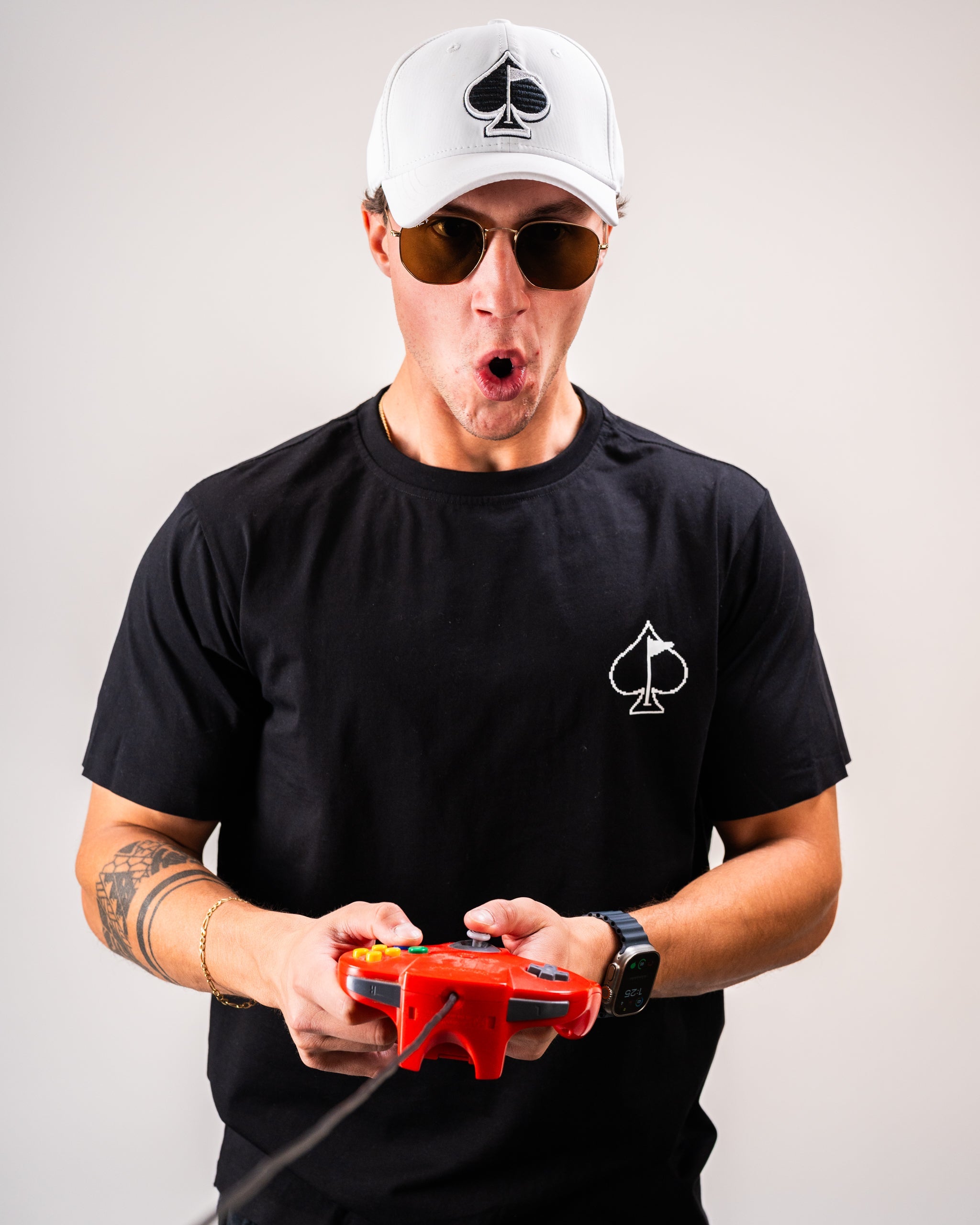Man in black t-shirt with spade logo playing with a red retro gaming controller, wearing a white cap and sunglasses, looking surprised.