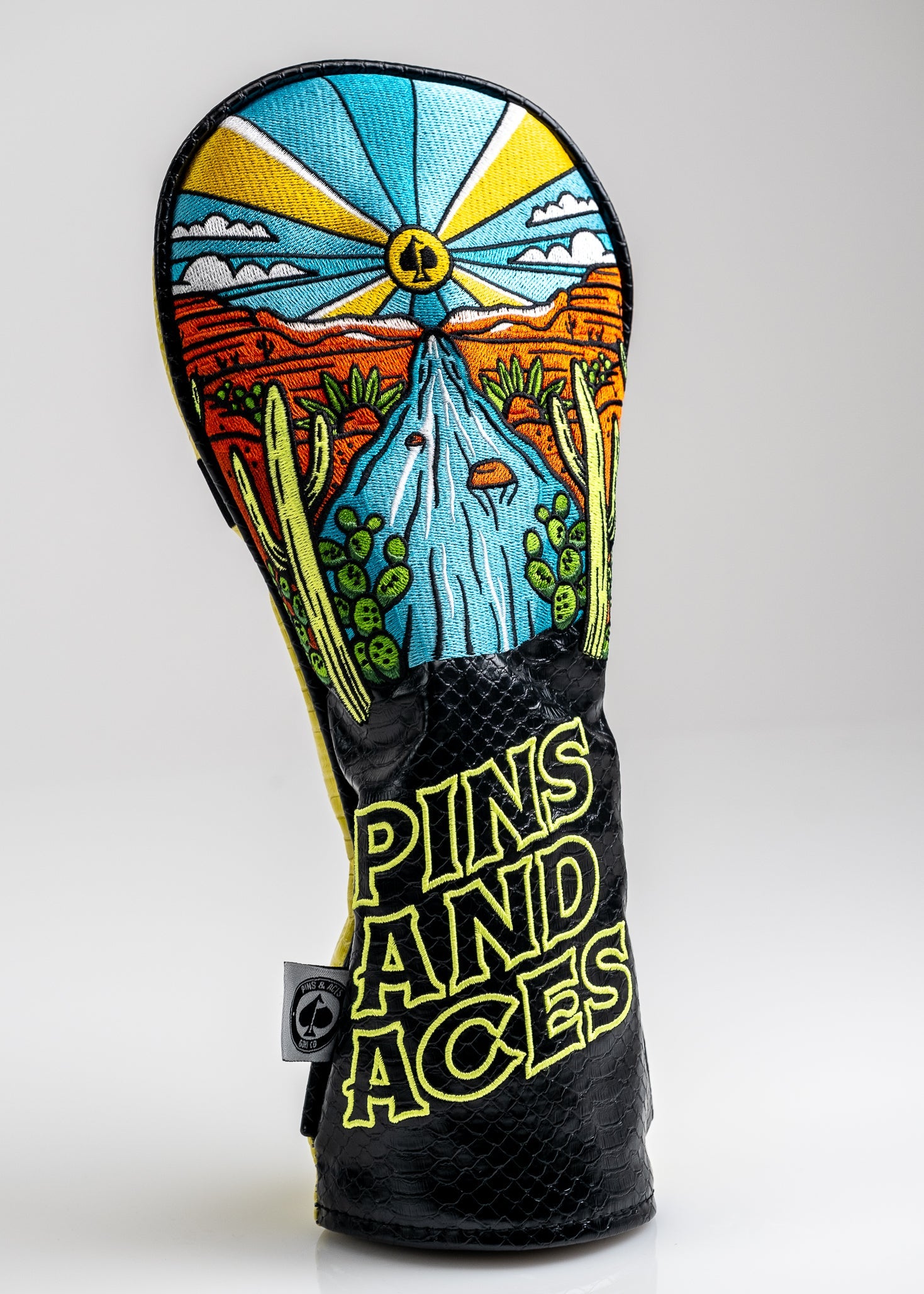 Colorful golf headcover featuring a desert scene with a water fountain, cacti, and a bright sun; bold "Pins and Aces" embr...