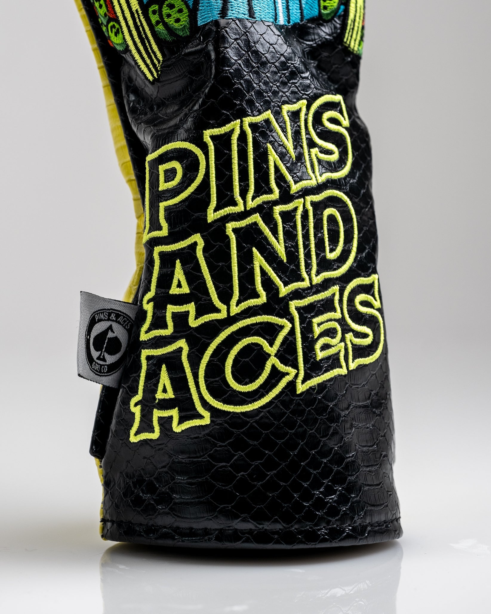 Bold black textured golf headcover with neon yellow embroidery reading "PINS AND ACES," featuring colorful, playful detail...