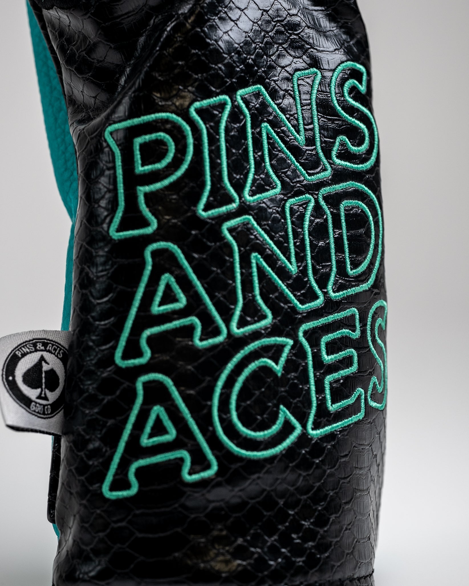 Black textured golf headcover with teal embroidered "PINS & ACES" logo, featuring bold, standout design for golfers who wa...
