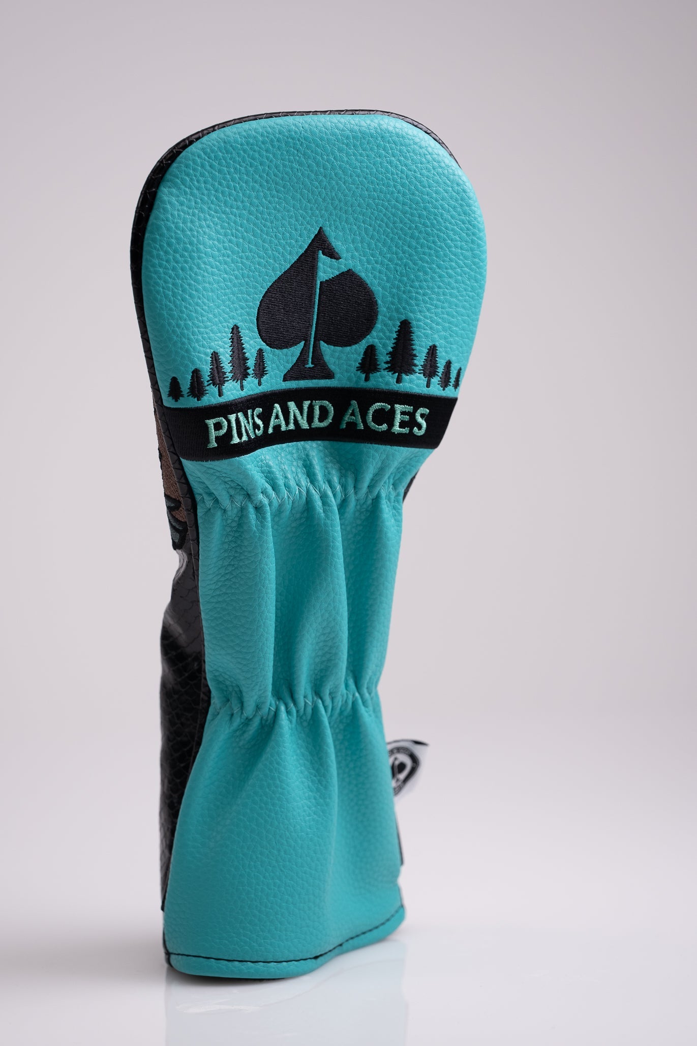 Colorful turquoise and black golf headcover with "Pins and Aces" logo, featuring a spade symbol and forest design, for bol...