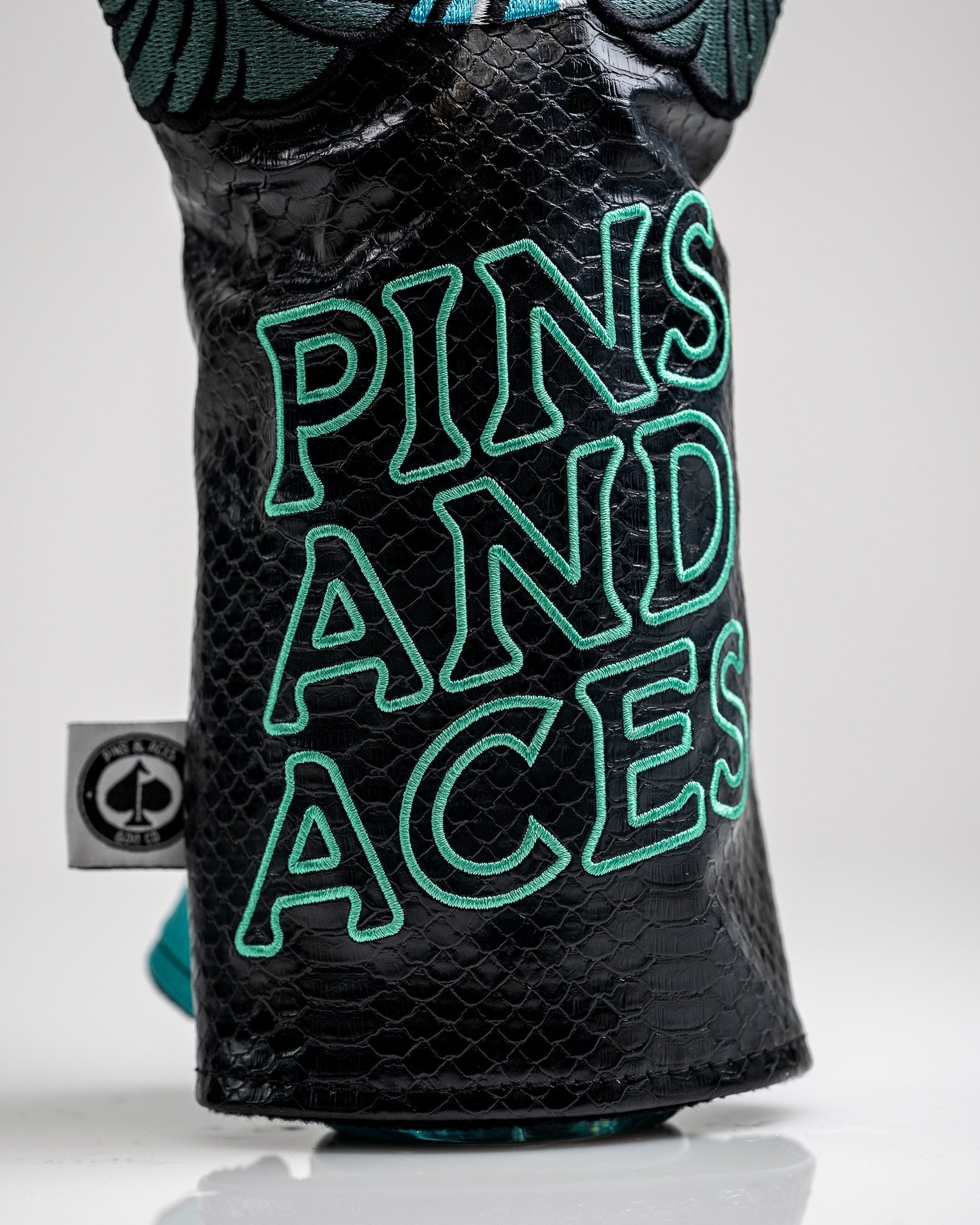 Black textured golf headcover with bold teal embroidered "PINS AND ACCES" text, featuring a snakeskin pattern and colorful...