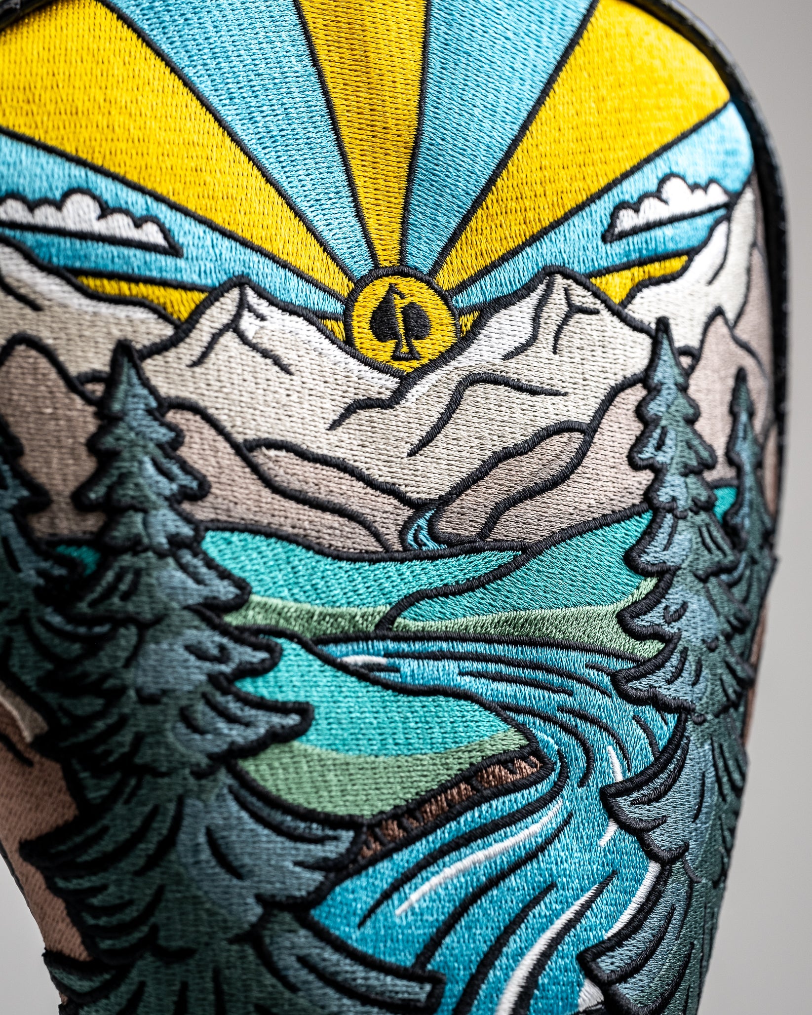 Colorful embroidered patch of a mountain landscape with a river, pine trees, and a sunburst in yellow, blue, green, and wh...