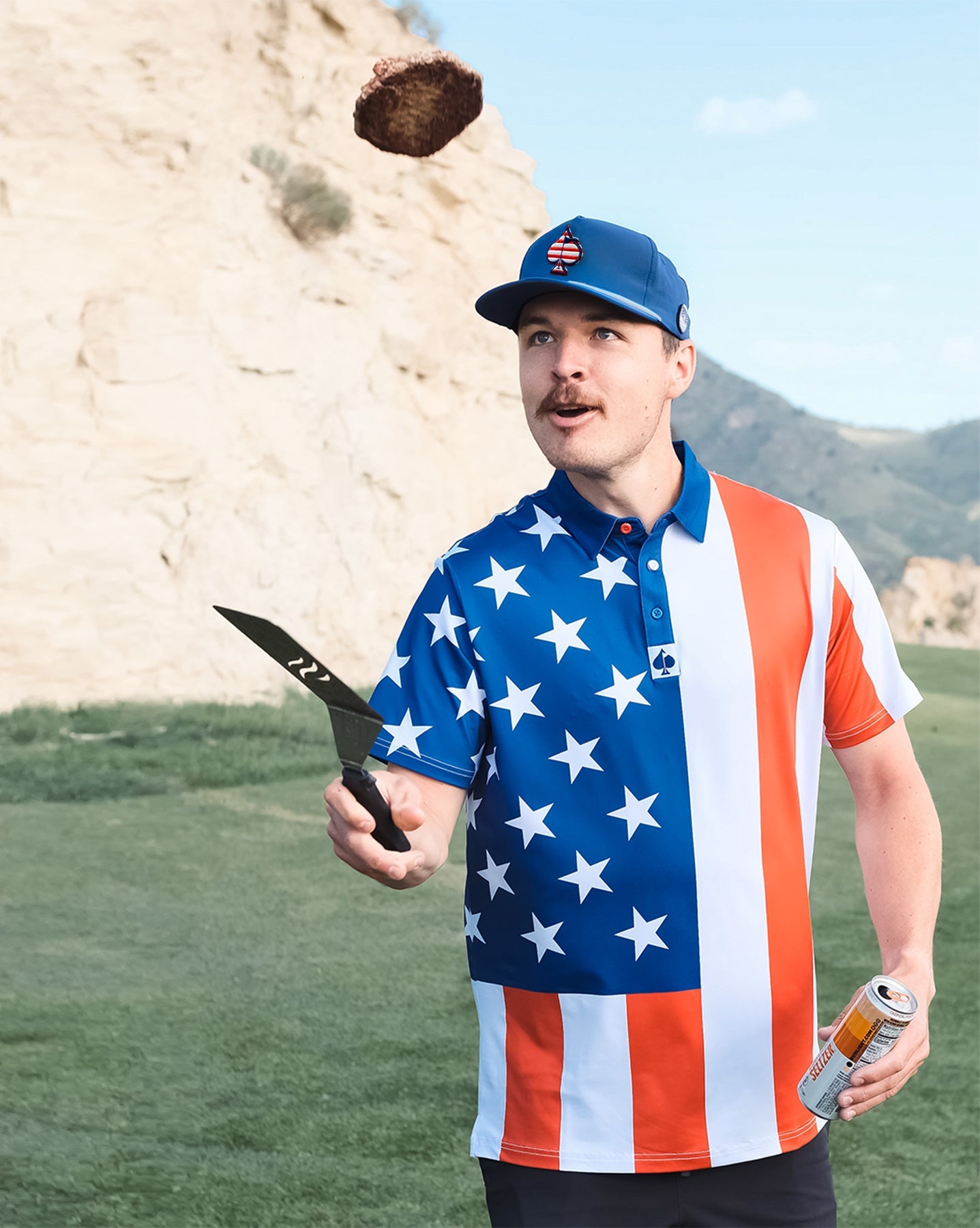 A man wearing a patriotic golf shirt and cap, holding a golf club and a can of beer outdoors with rocky cliffs in the background.