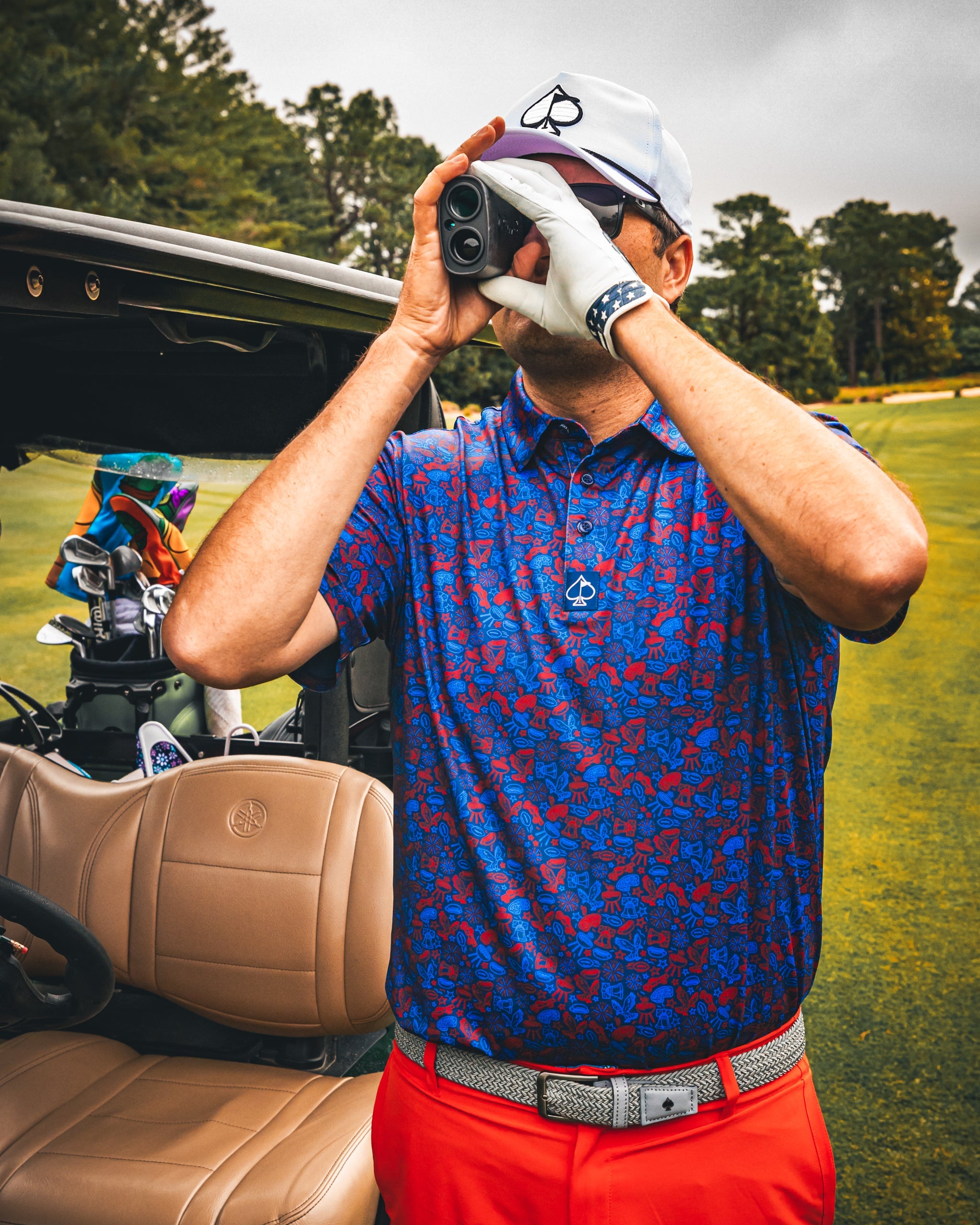 A golfer in a navy polo shirt with a doodle pattern uses a rangefinder on the course, wearing white gloves and red pants.