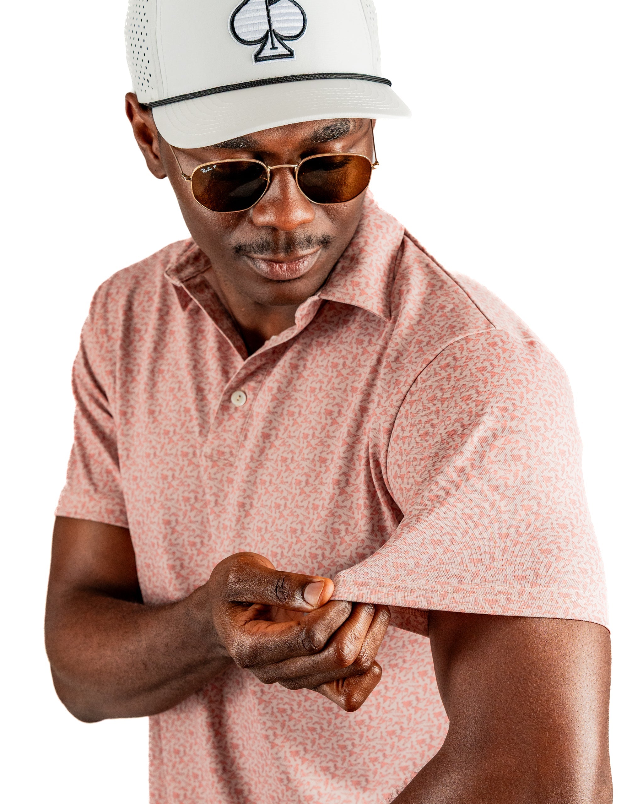A man in a pink patterned polo shirt adjusts the sleeve while wearing sunglasses and a white cap.