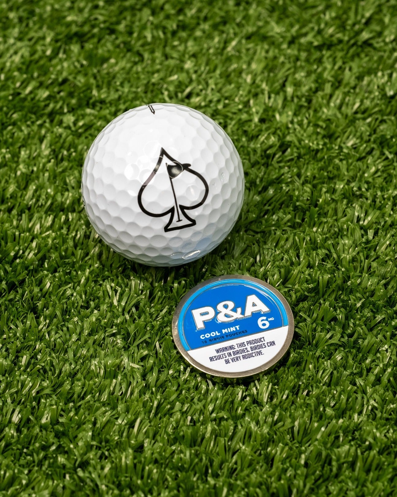 Cool Mint ball marker next to a golf ball with a black spade logo, set on green turf. Perfect for stylish golfers.