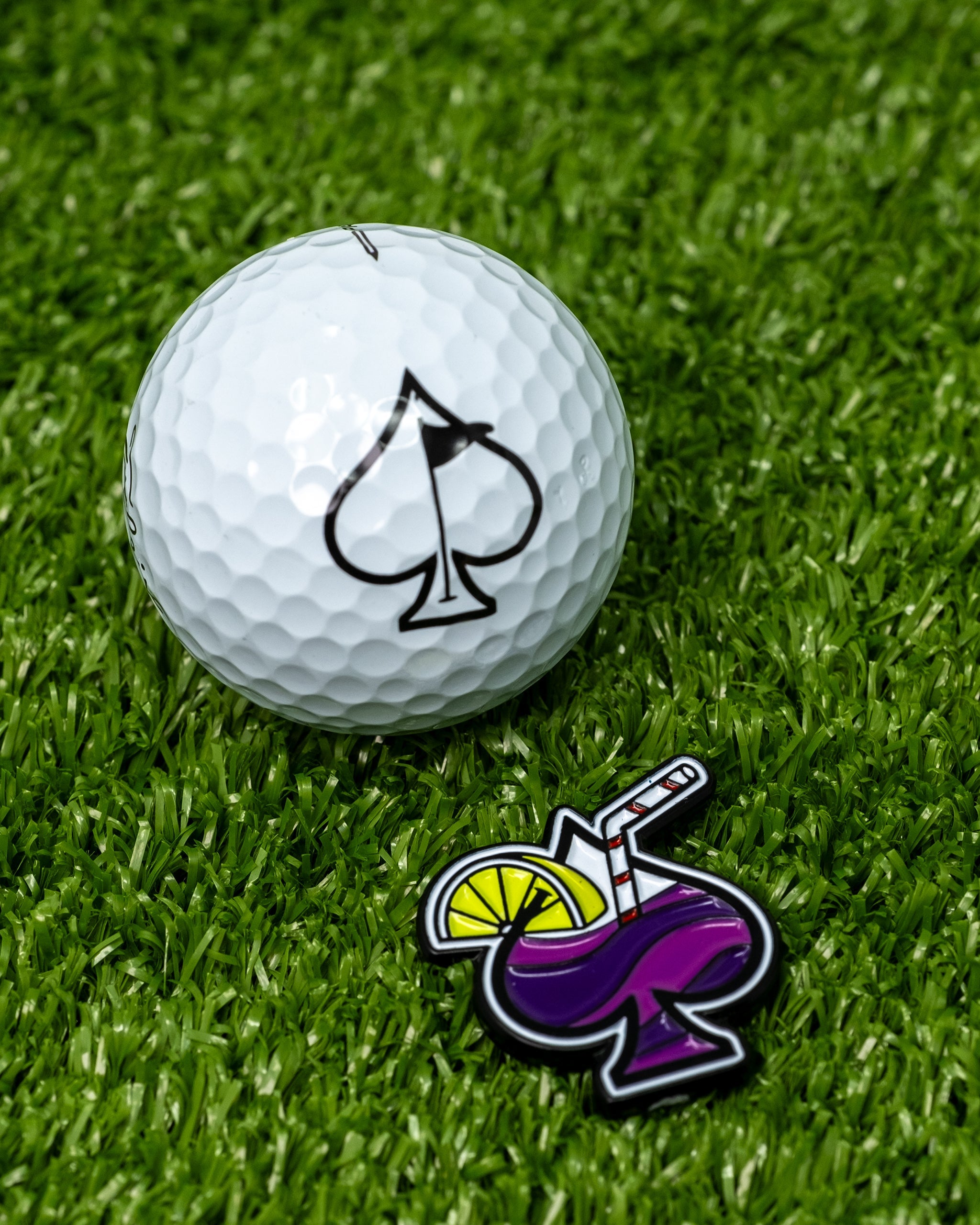 Transfusion Spade Ball Marker next to a white golf ball featuring a spade logo, set on a green grass background.