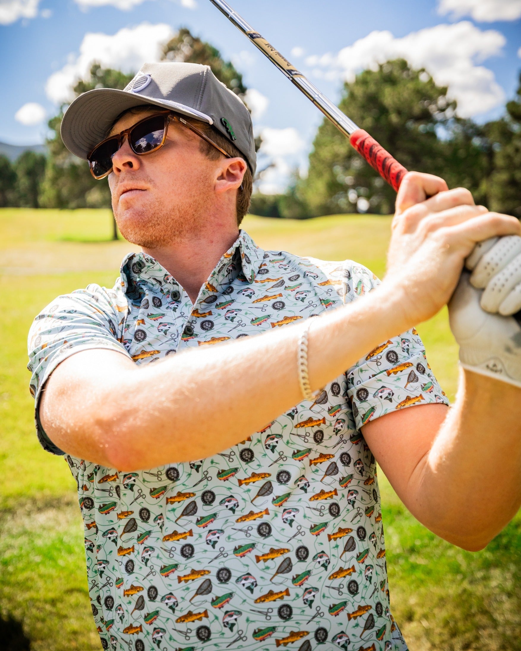 A golfer in a patterned shirt featuring fishing and golf motifs, preparing to swing on a sunny course.