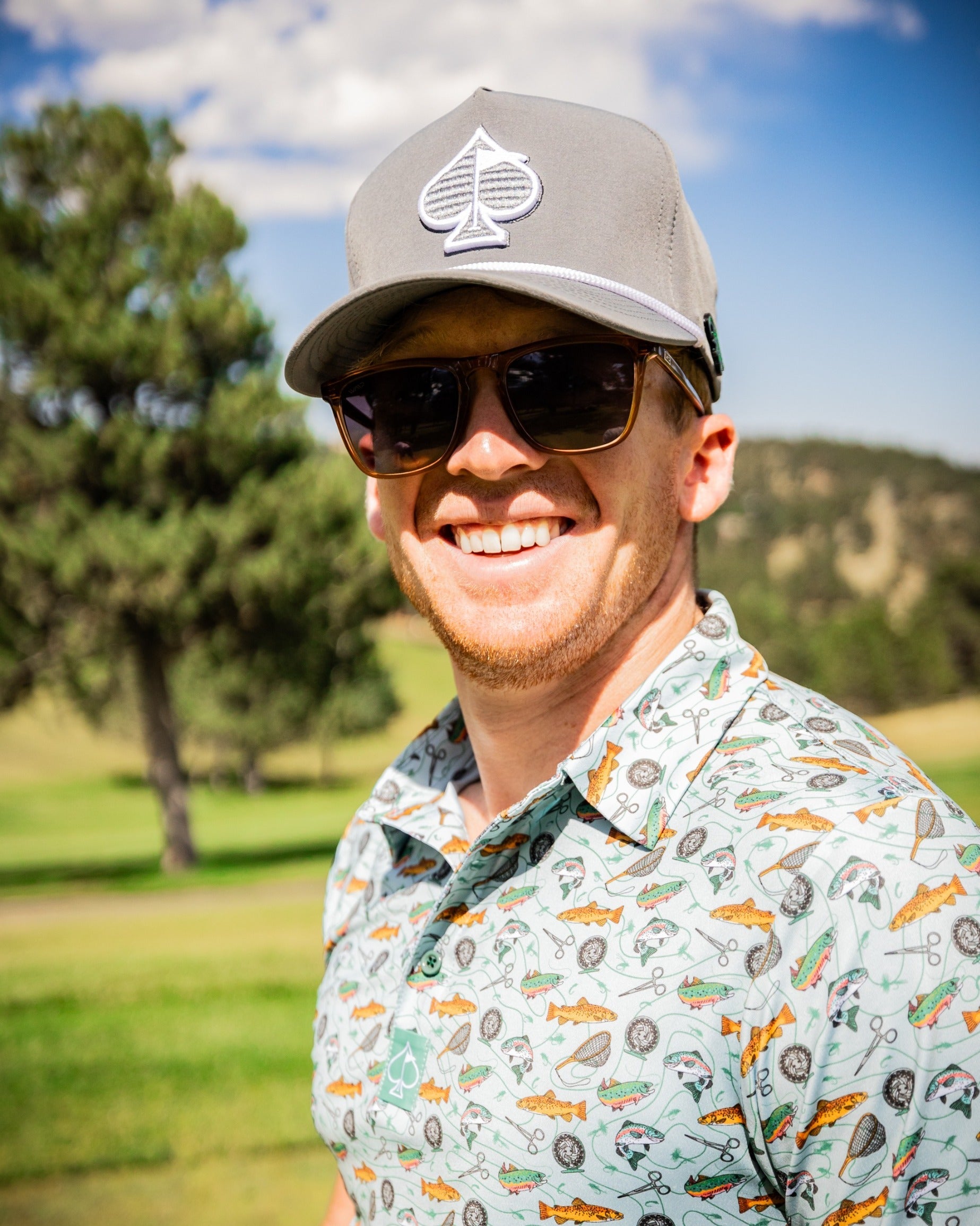 Man wearing a gray cap and a colorful fishing-themed polo shirt, smiling on a golf course with trees in the background.