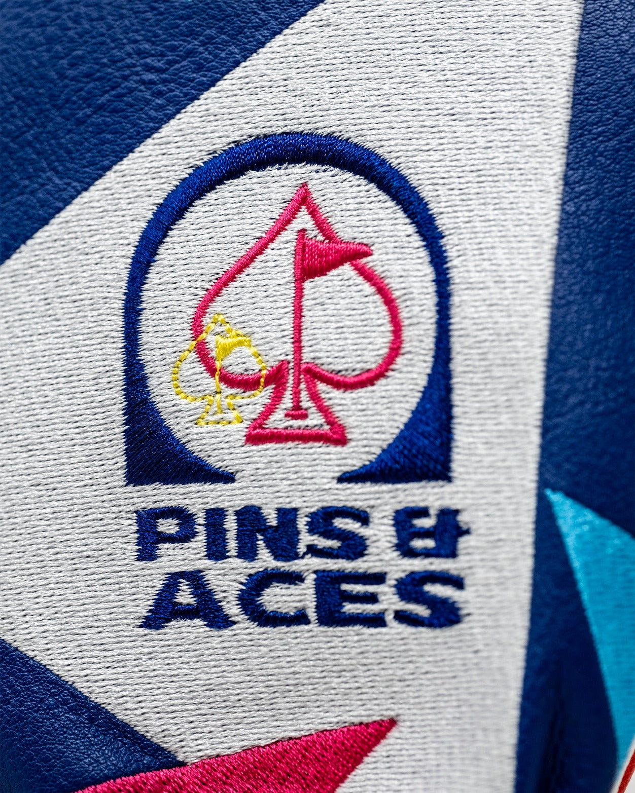Embroidered logo of Pins and Aces on a vibrant fairway cover, featuring colorful spade and club designs.