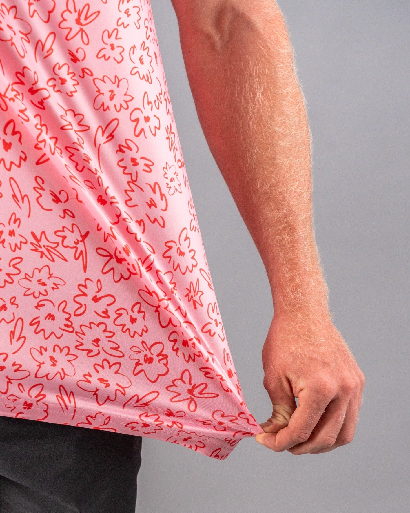A close-up of a man holding the hem of a pink golf shirt featuring a playful flower doodle pattern in red.