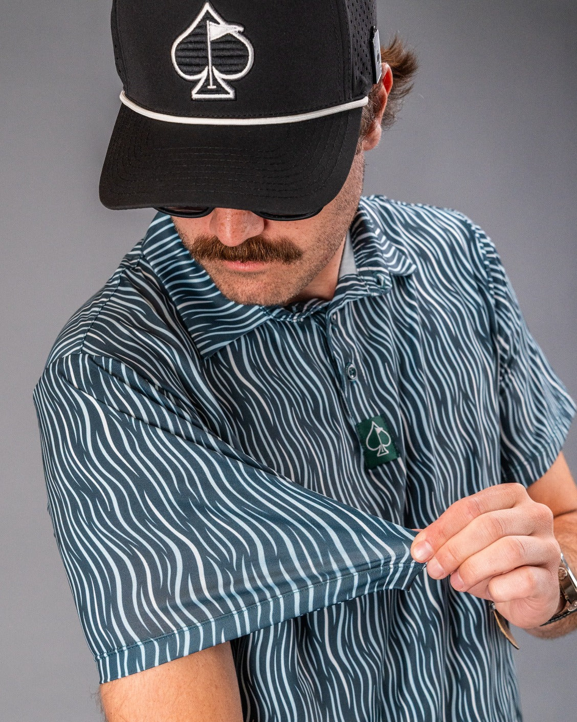 Close-up of a man wearing a black golf cap with a white spade logo, striped golf shirt, and standing against a neutral bac...