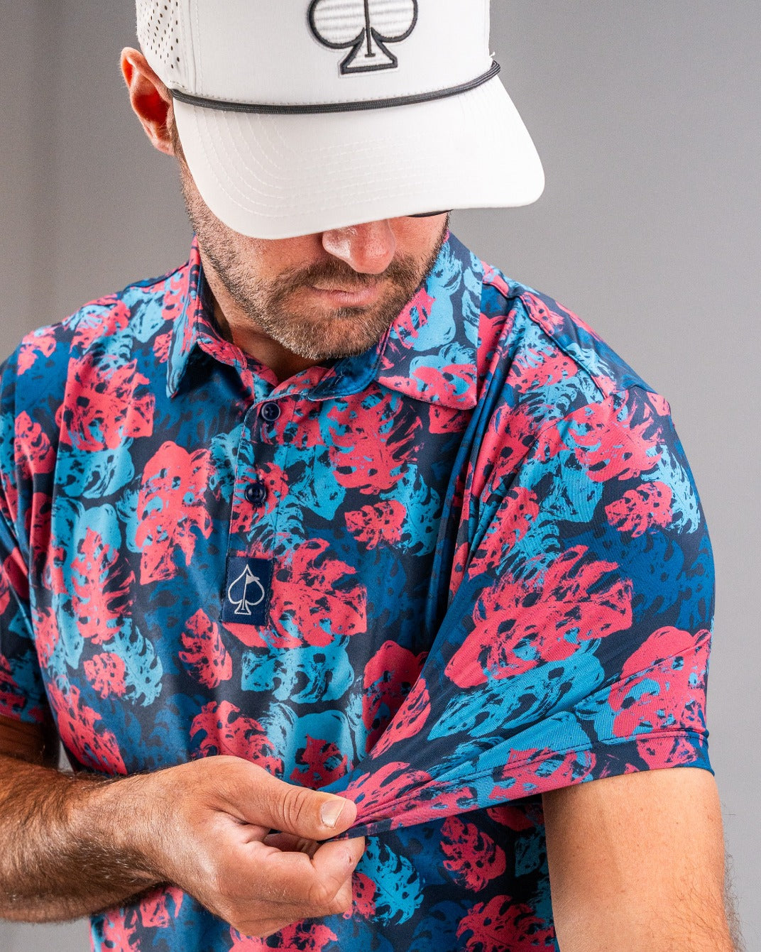 Close-up of a golfer wearing a vibrant blue and pink floral patterned polo shirt with a white cap featuring a black spade ...
