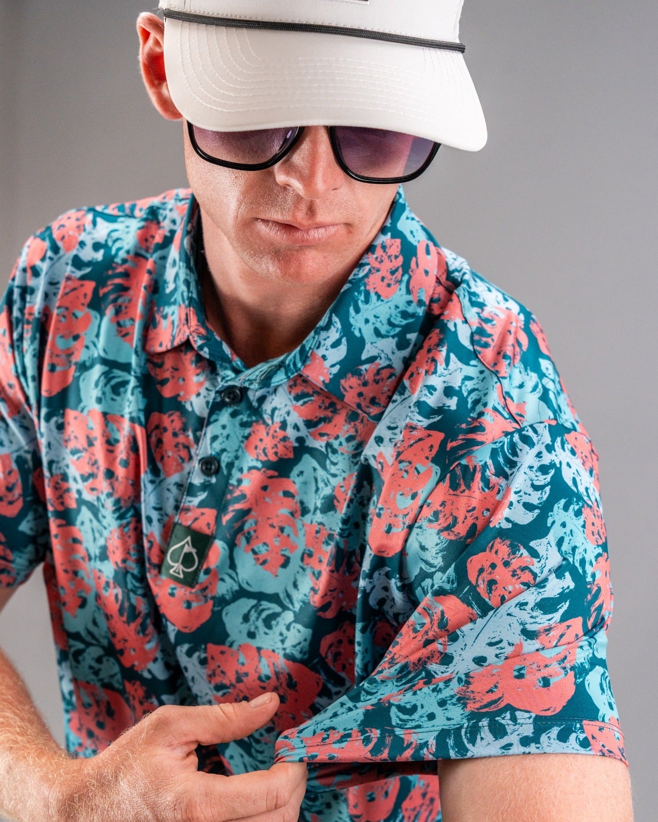 Man wearing a bold, colorful floral-patterned golf shirt in teal, coral, and black, with a white golf cap and dark sunglas...