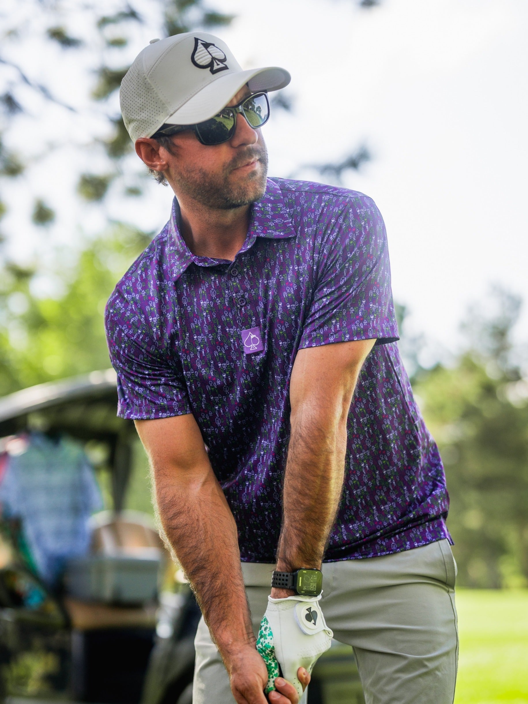 Male golfer wearing a purple patterned polo, white cap with spade logo, sunglasses, and a golf glove, on a sunny course.