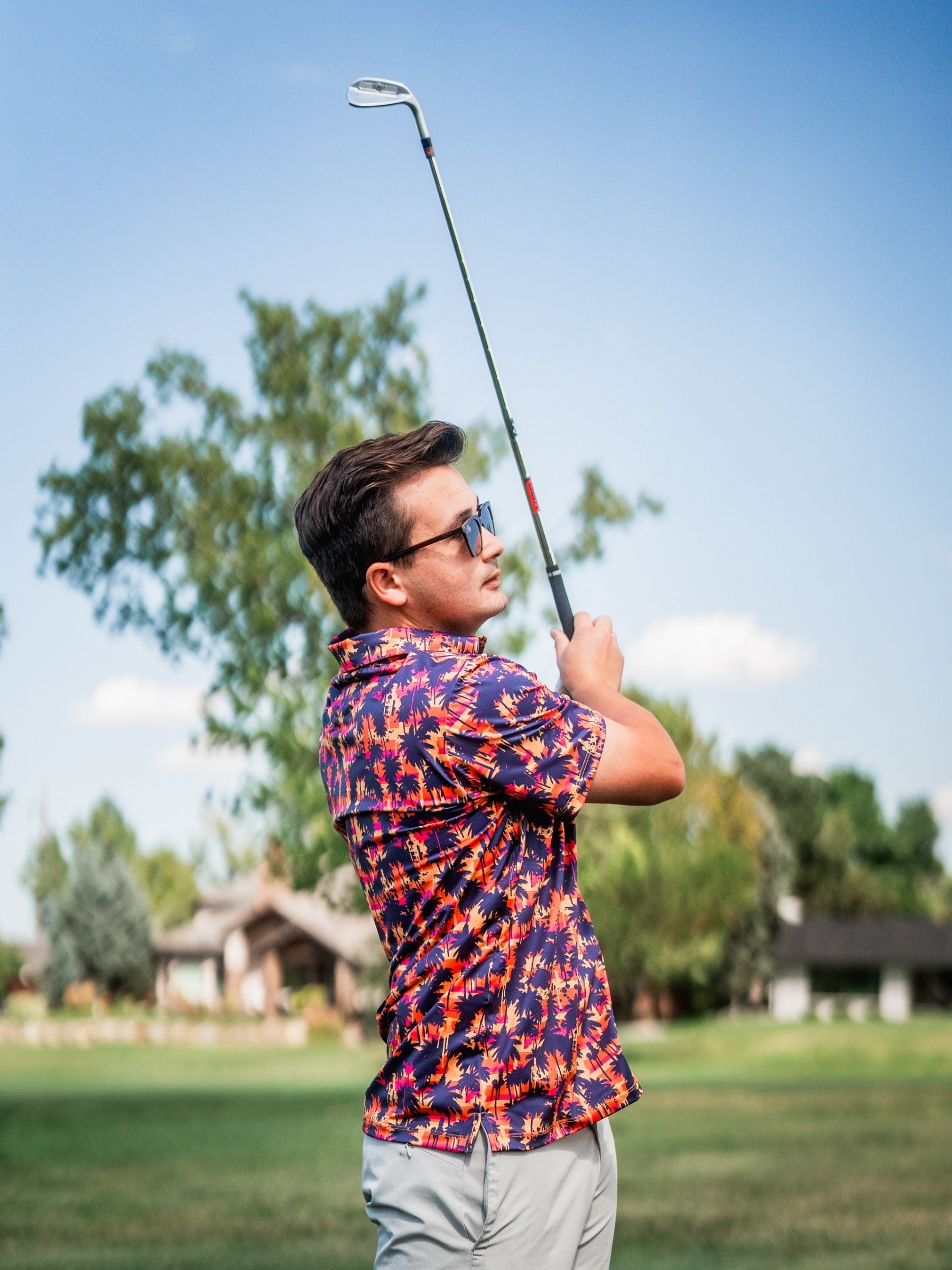 Young man with dark hair and sunglasses, wearing a colorful tropical golf shirt, swings a golf club on a sunny course.