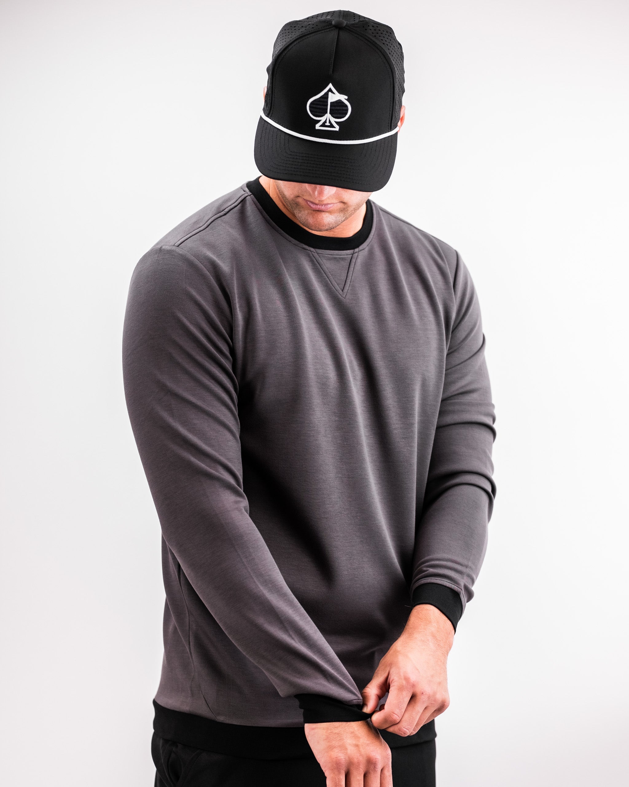 Man wearing a slate grey Player Preferred™ crewneck sweatshirt, adjusting the sleeve, with a black cap featuring a white spade logo.