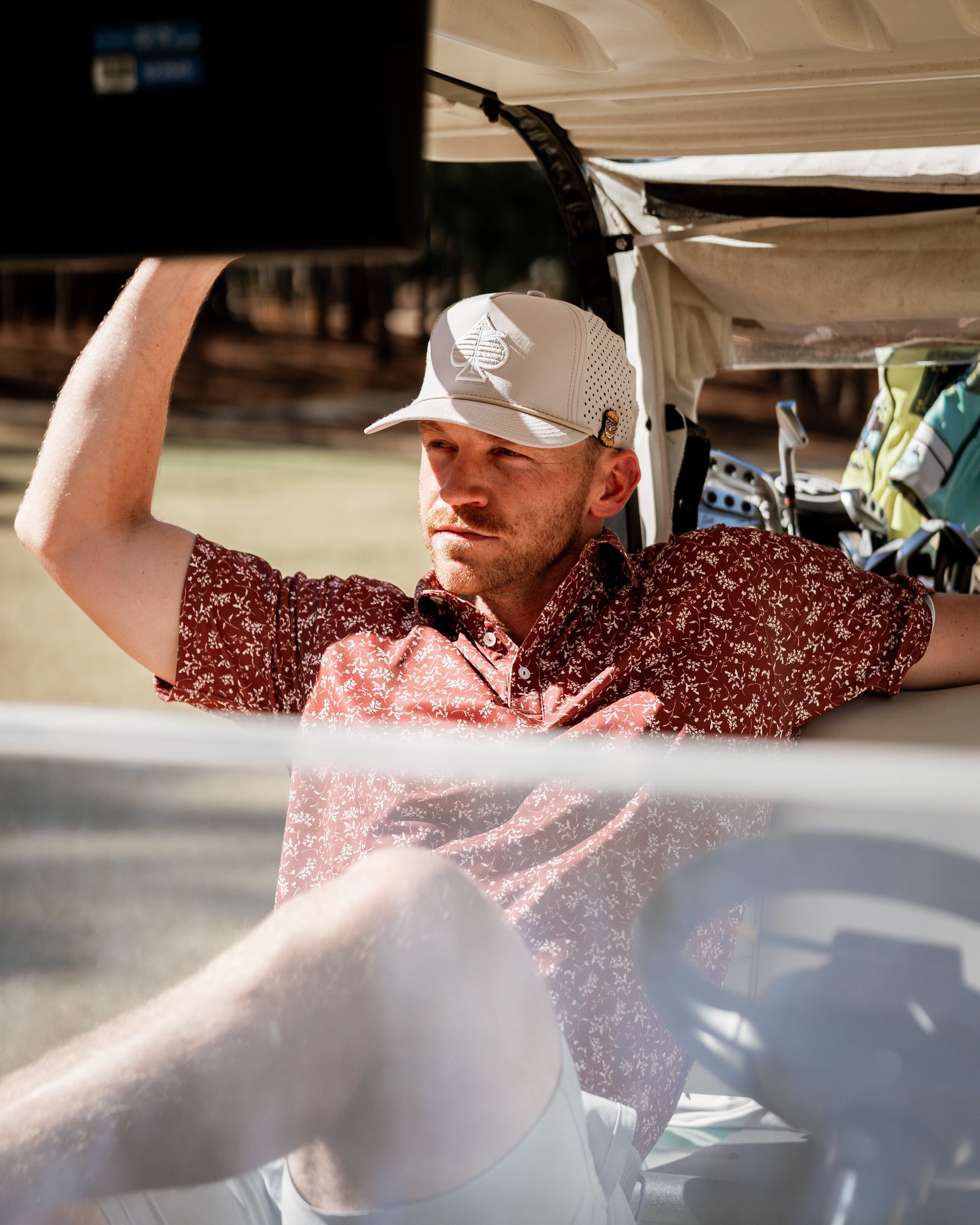 Man relaxing in a golf cart, wearing a floral-patterned shirt and a cap, enjoying a sunny day on the golf course.