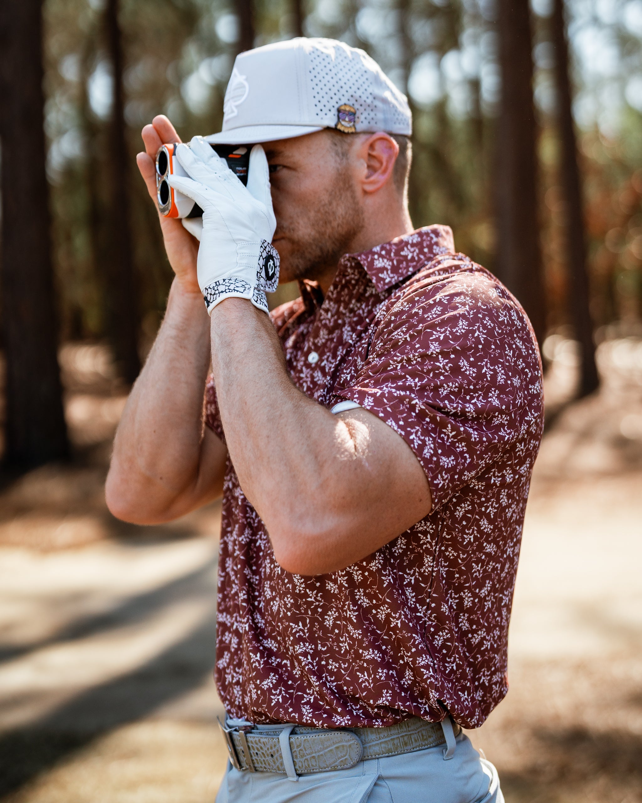 Man wearing a maroon floral golf shirt, holding a rangefinder on a golf course surrounded by trees.
