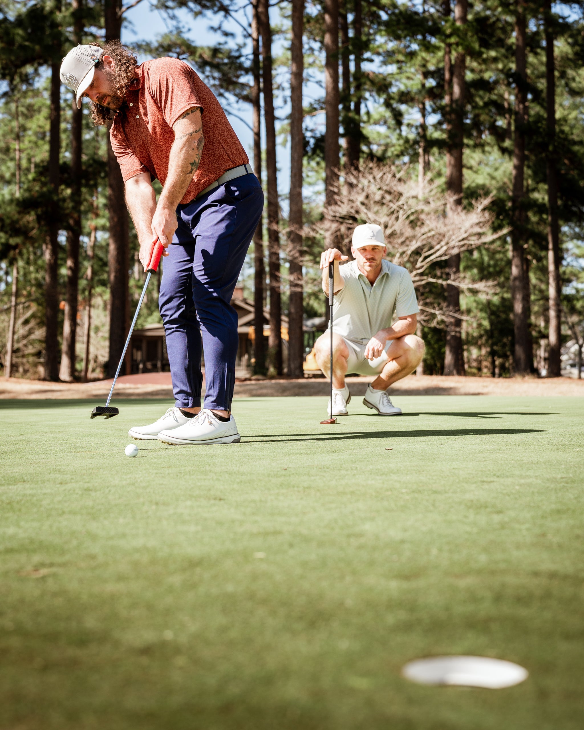 Two golfers on a green, one putting while the other observes. Both wear stylish golf apparel in a wooded setting.