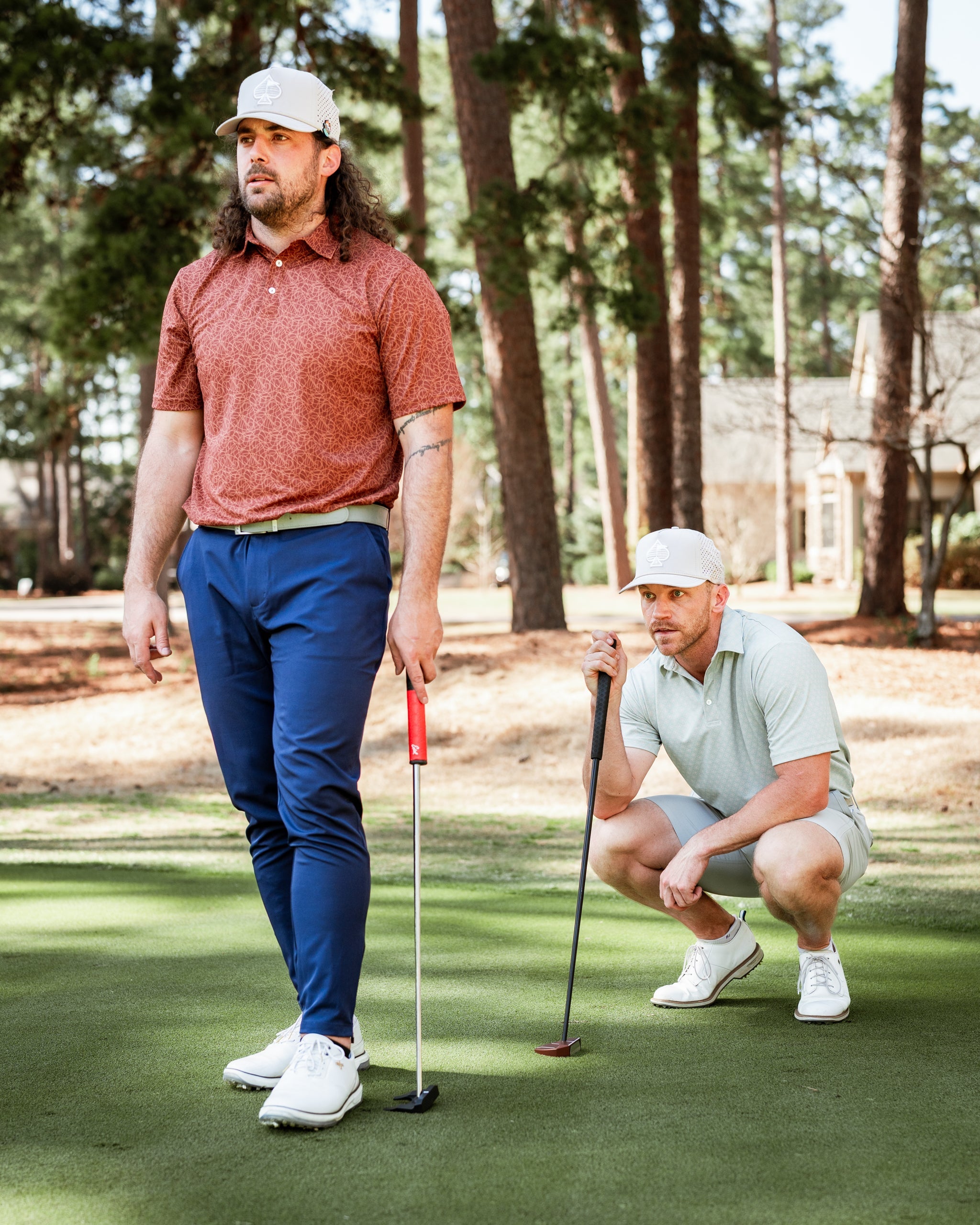 Two golfers on a course wearing stylish golf apparel, one preparing to putt while the other observes, surrounded by trees.