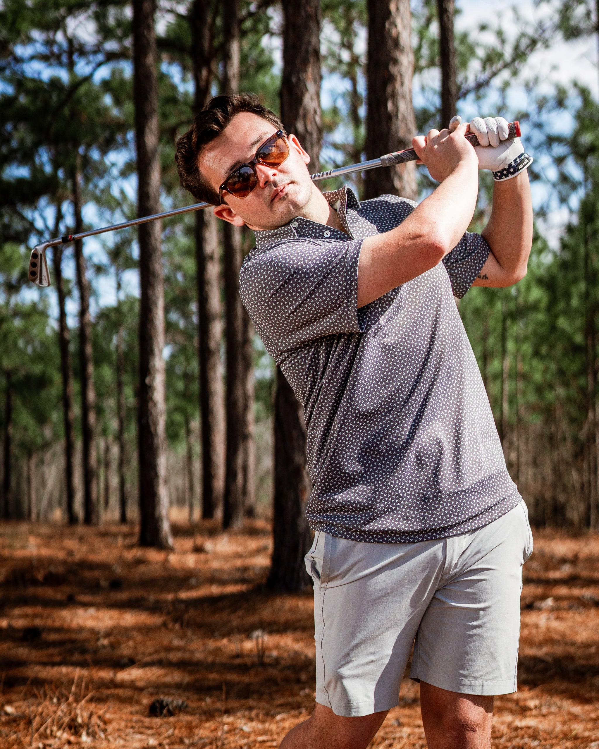 A golfer swings a club in a forested area, wearing a stylish polka dot shirt and sunglasses, showcasing the Jade Drops apparel.