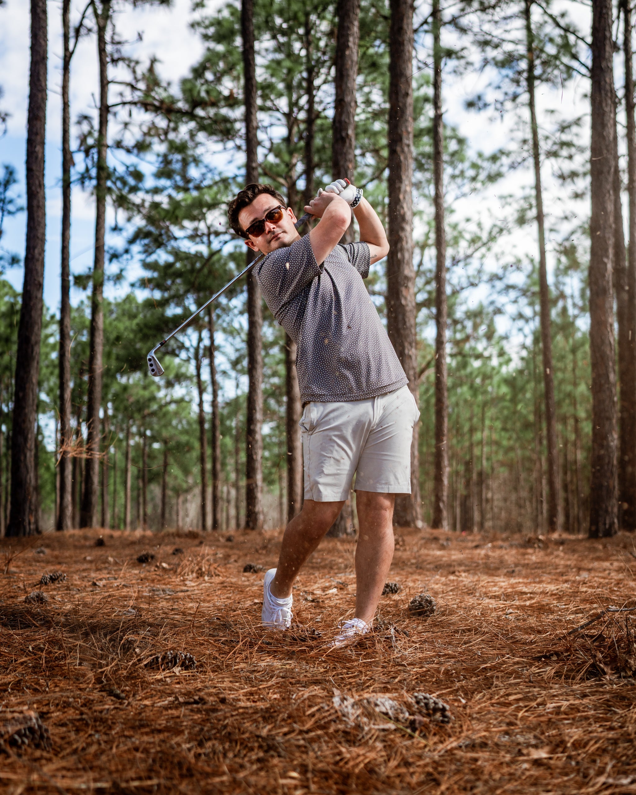A golfer swings a club in a forested area, wearing a stylish gray polo and light shorts, showcasing confidence and style.