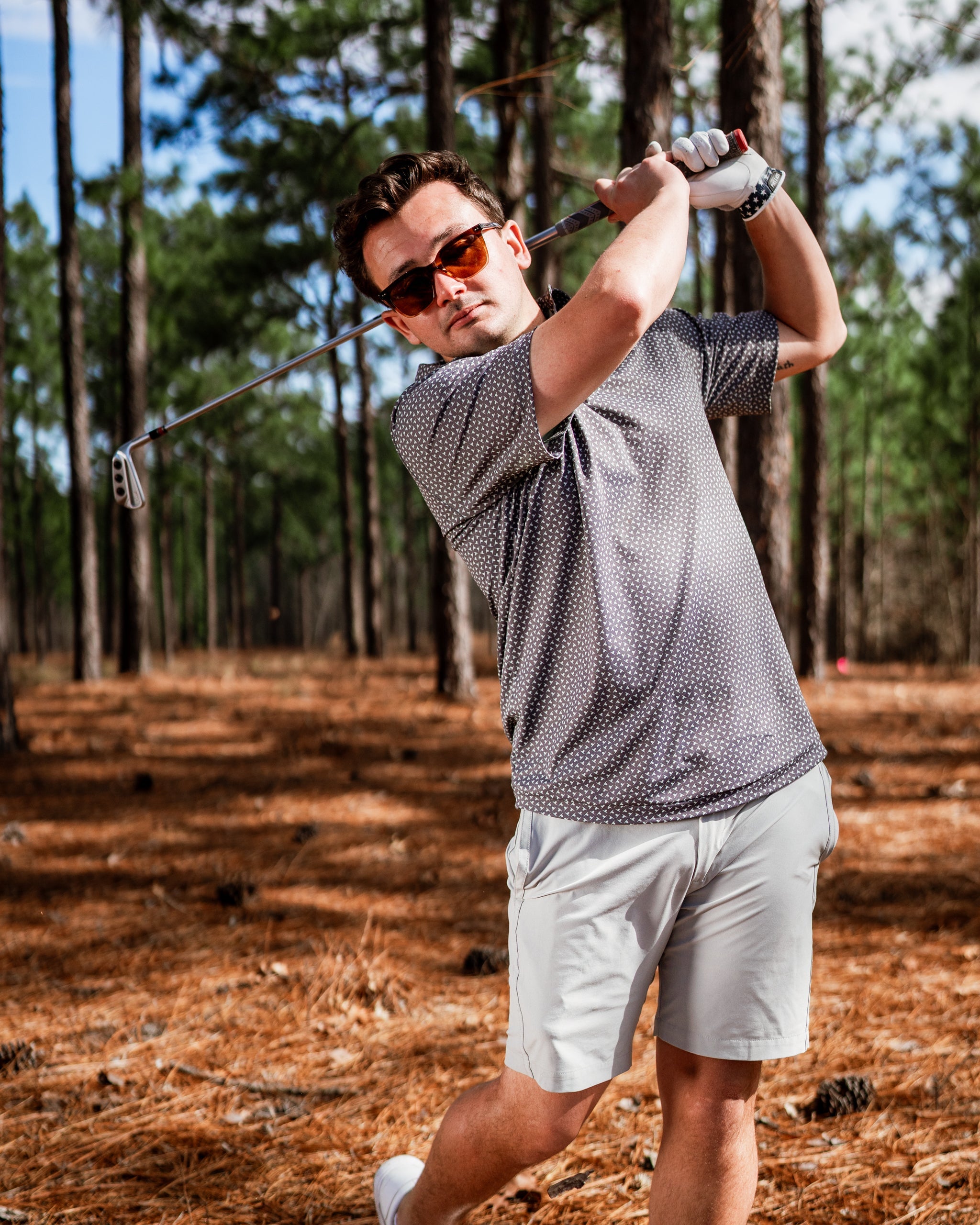 A golfer swings a club in a forested area, wearing a stylish patterned shirt and sunglasses, showcasing bold golf fashion.