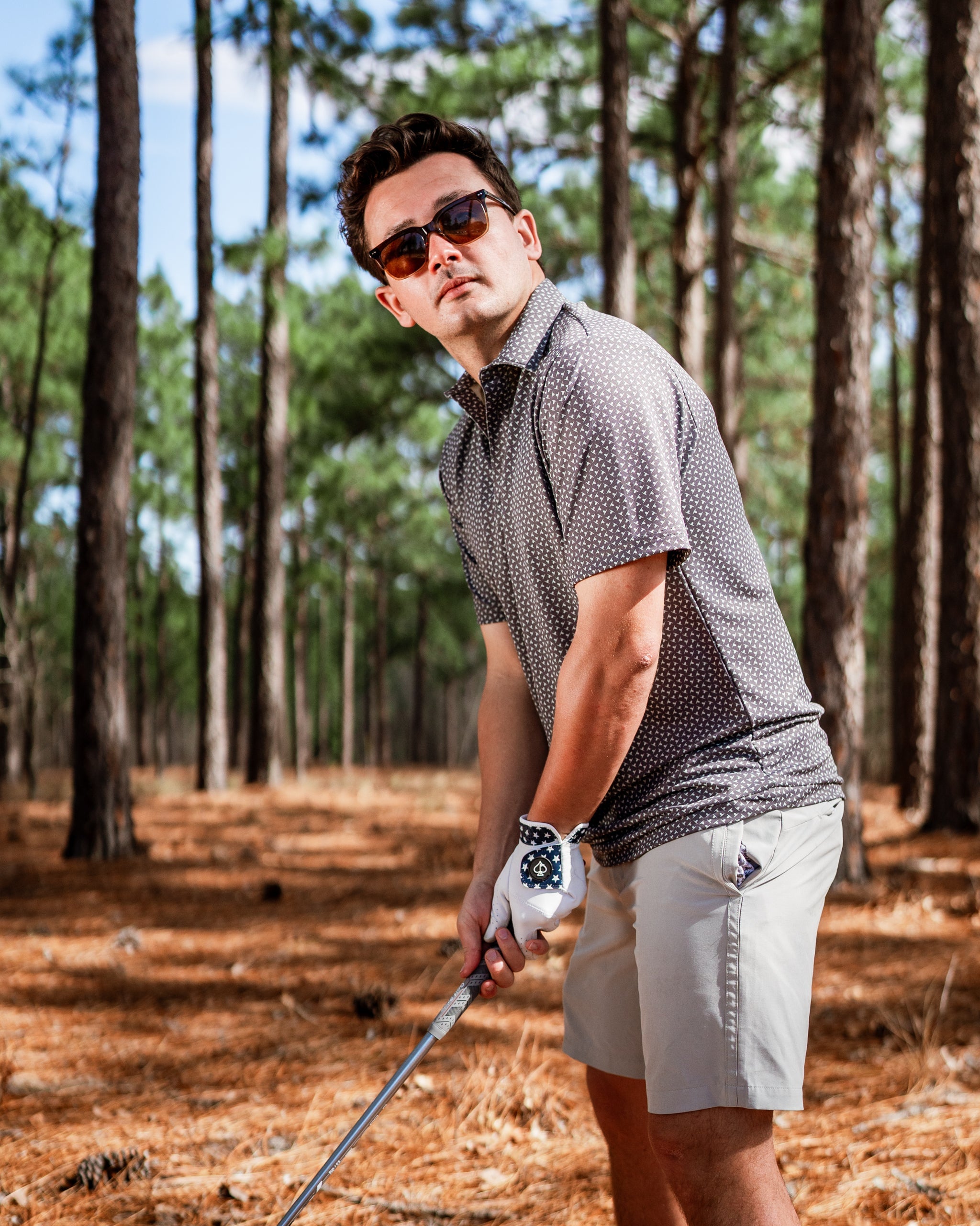 A golfer wearing a stylish patterned polo and shorts prepares to swing in a pine forest setting.