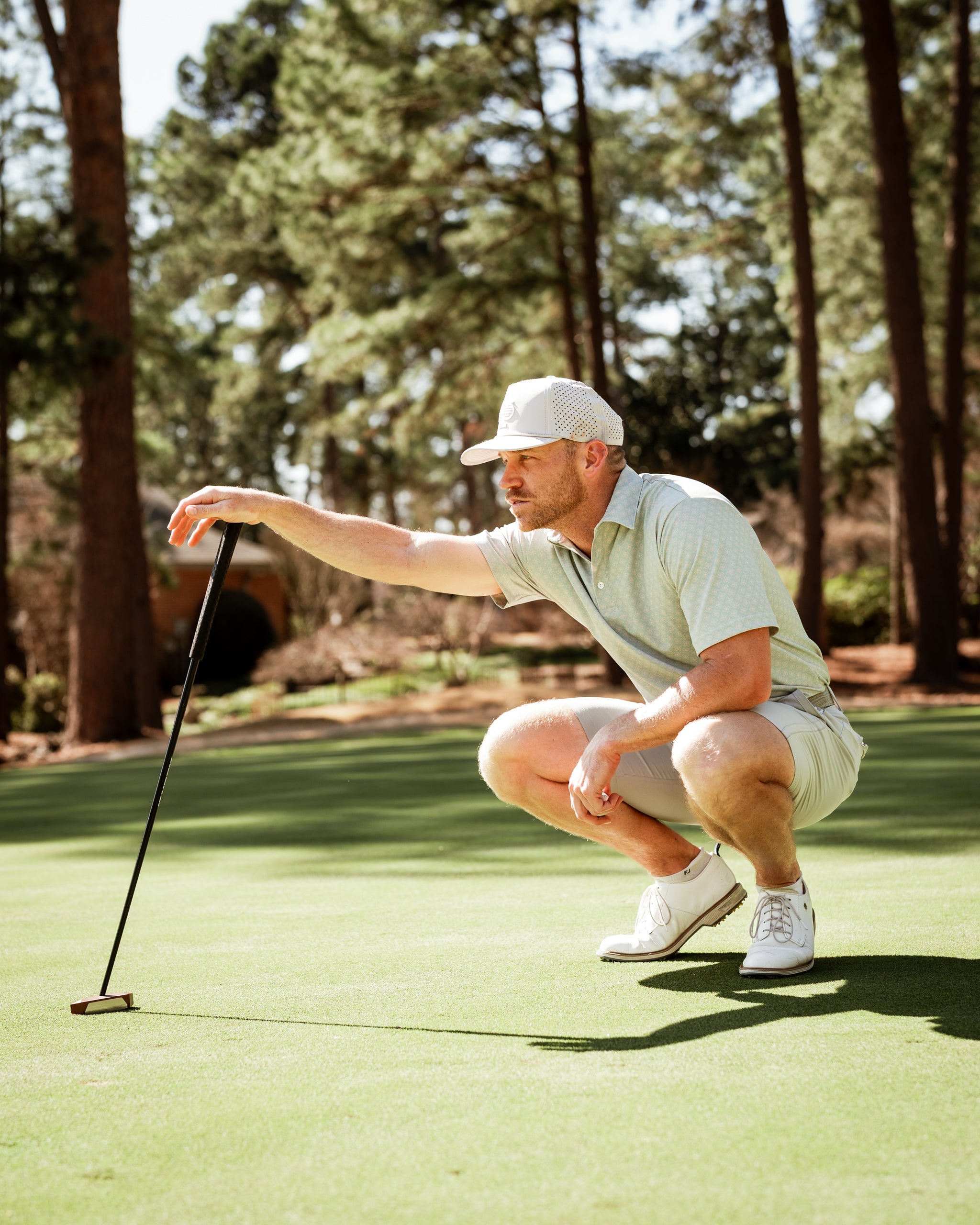 A golfer in a light green polo and shorts crouches on the putting green, focusing intently on his putt.