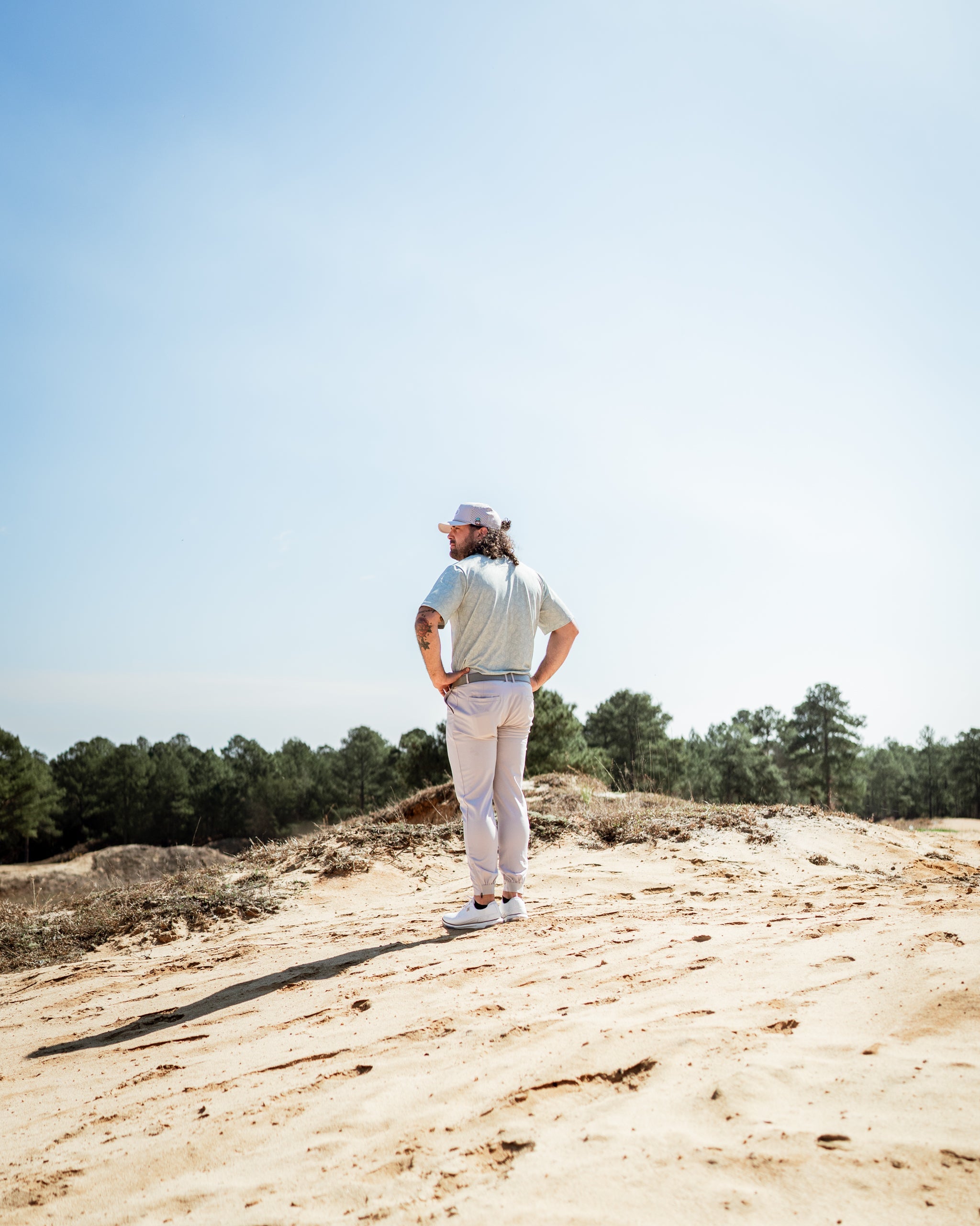 A golfer stands on sandy terrain, wearing a light-colored shirt and pants, with trees in the background under a clear sky.