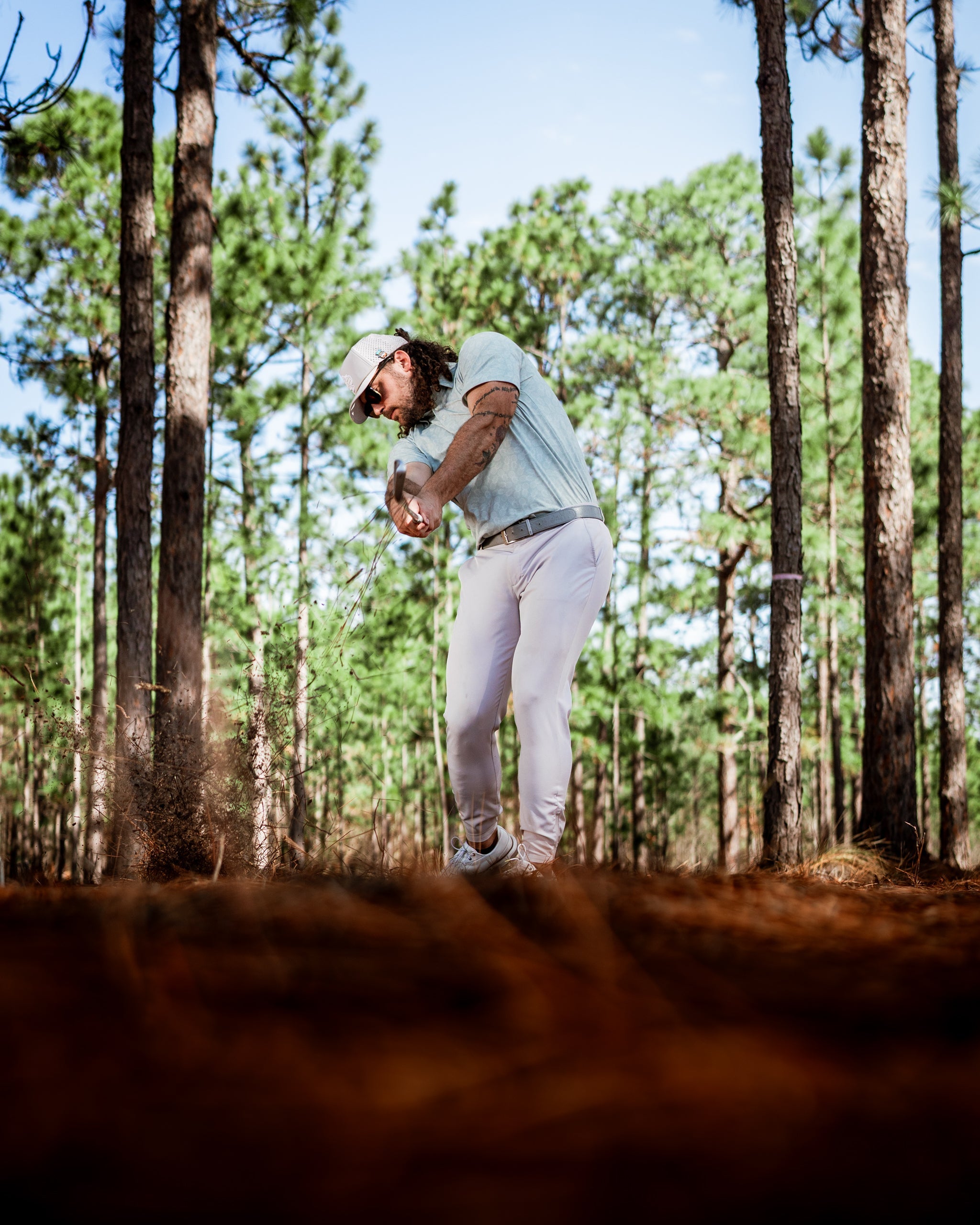 Golfer in a light blue shirt and white pants swings a club in a forested area, surrounded by tall pine trees.