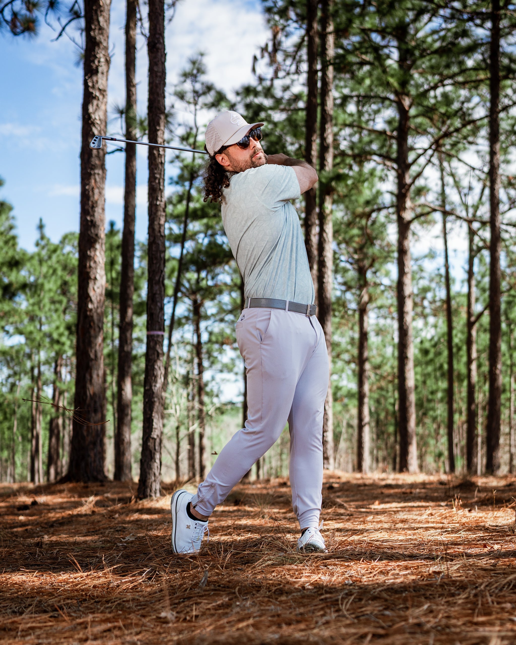 Golfer in light gray apparel swings a club in a pine forest, showcasing a stylish and bold golf look.