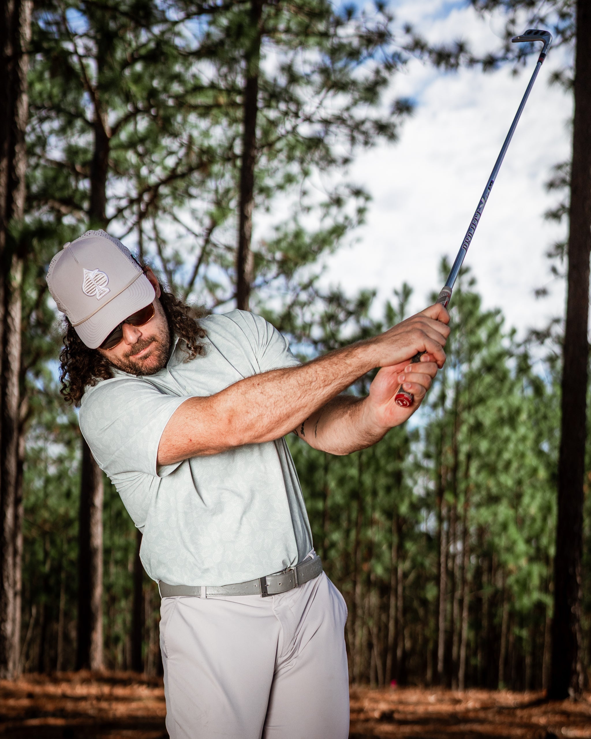 Golfer in a light green shirt and cap swings a golf club in a pine forest, showcasing bold golf apparel.