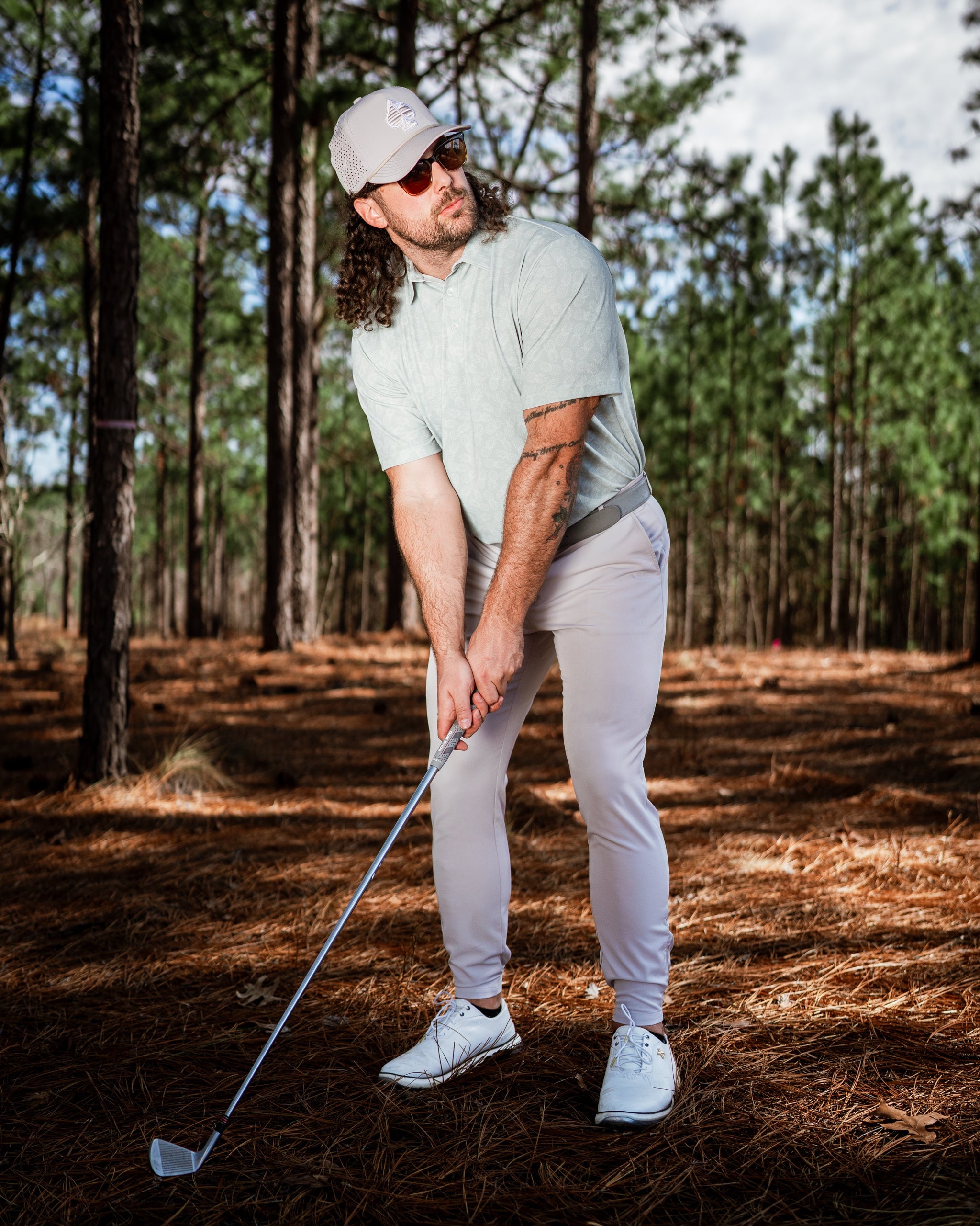 A golfer in a light green polo and white pants prepares to swing a club in a pine forest setting.