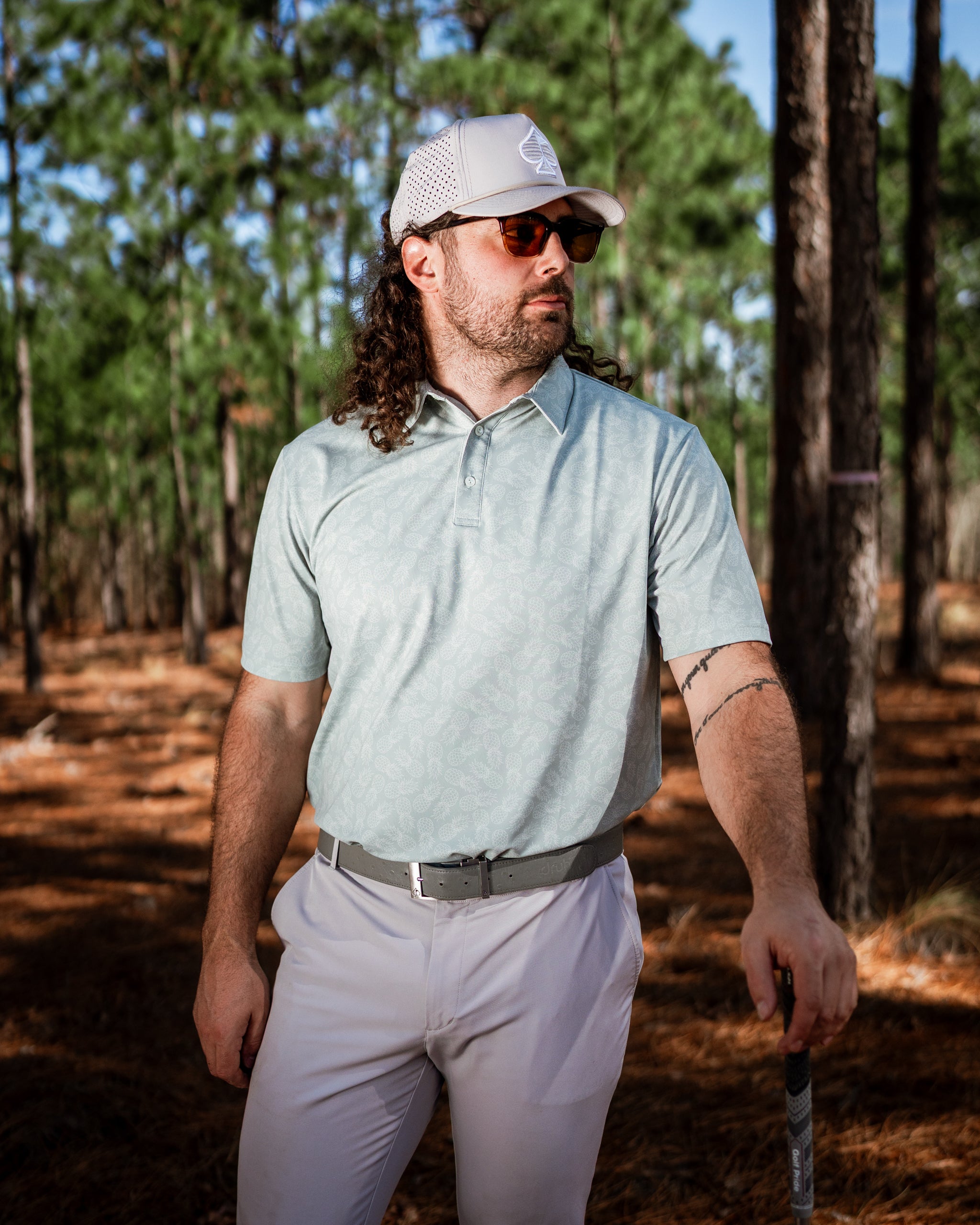 Man wearing a light green polo with a pineapple pattern, standing confidently in a wooded golf course setting.