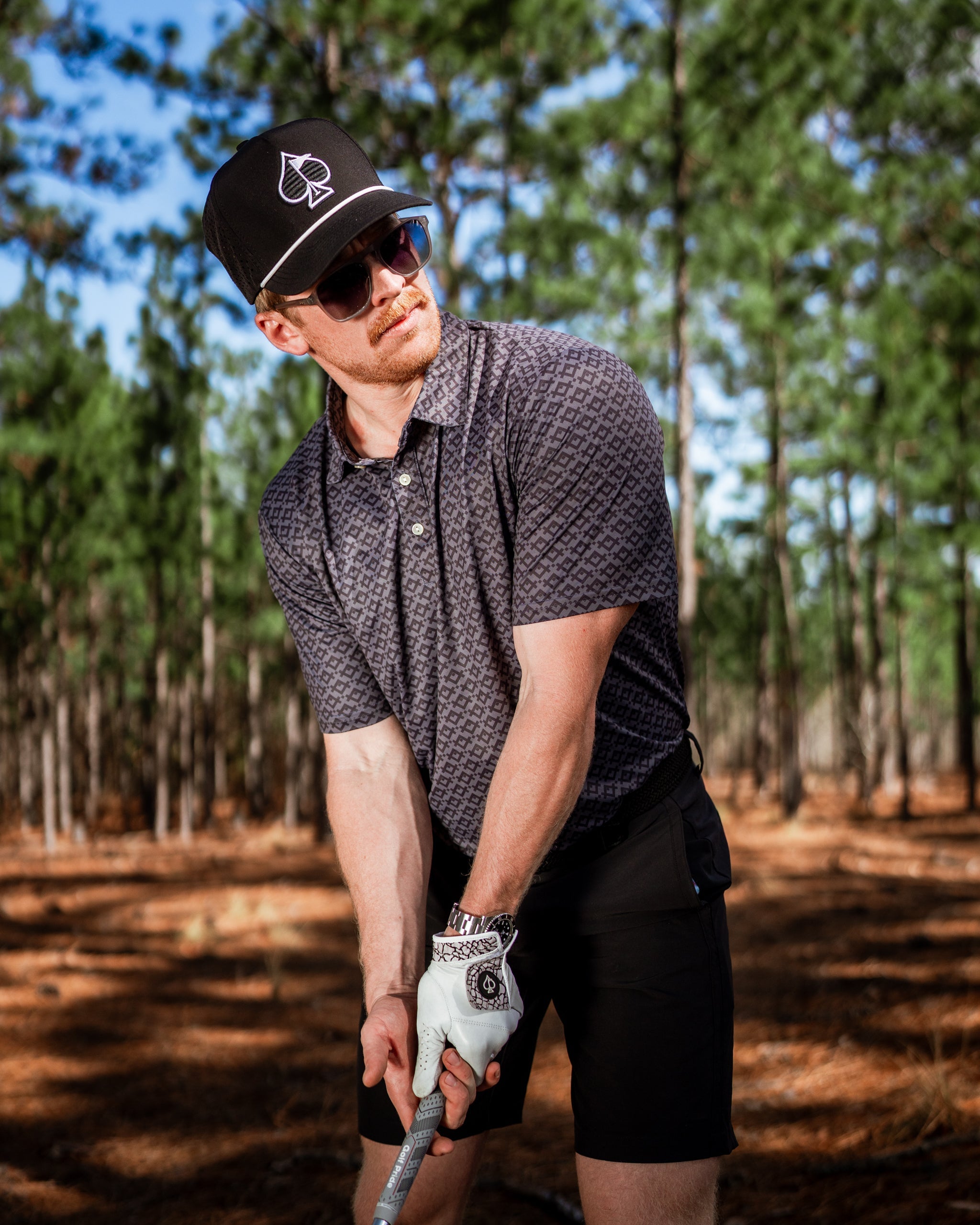 Stylish golfer wearing a Slate Mosaic polo and cap, preparing to swing in a wooded golf course setting.