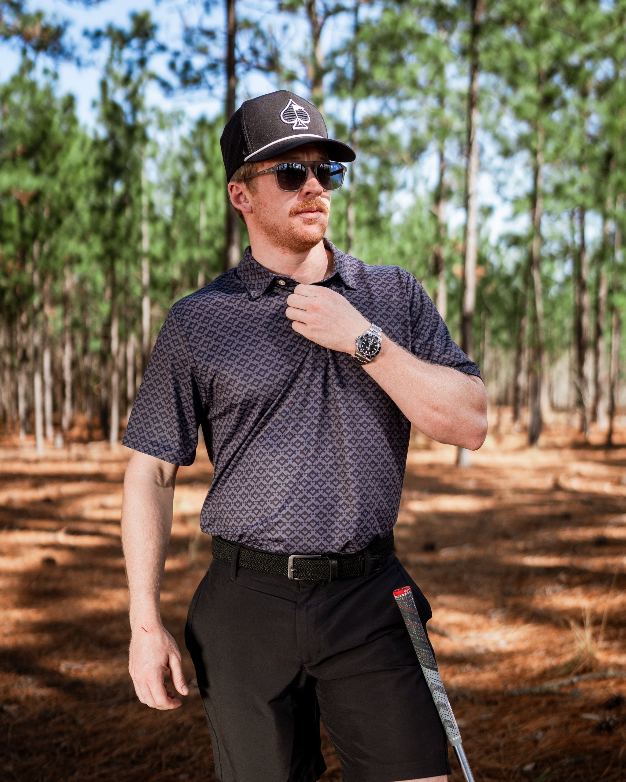 Man wearing a Slate Mosaic golf polo shirt, sunglasses, and a cap, standing in a wooded area with pine trees.