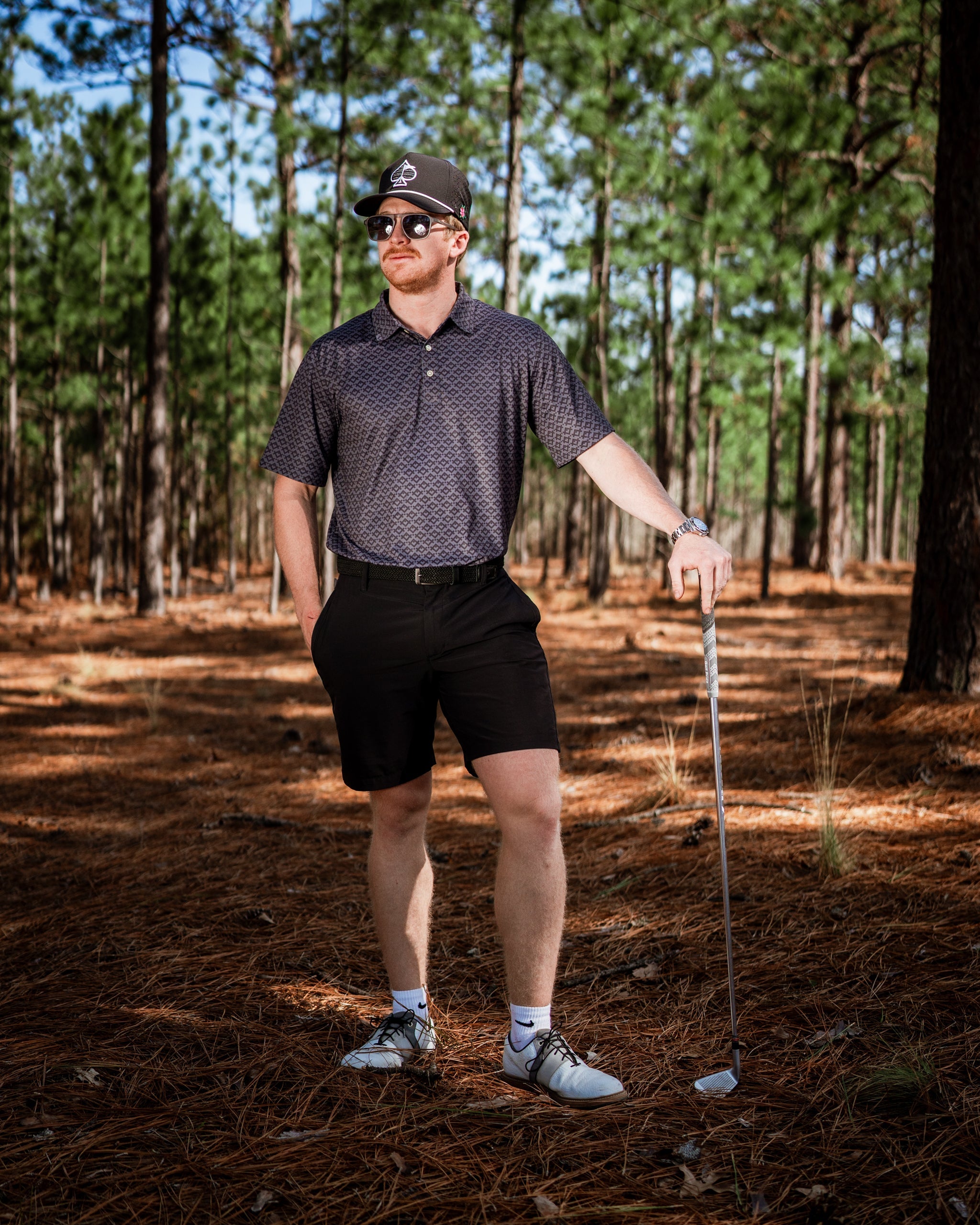 A golfer stands confidently in a forest wearing a slate mosaic polo and black shorts, holding a golf club.