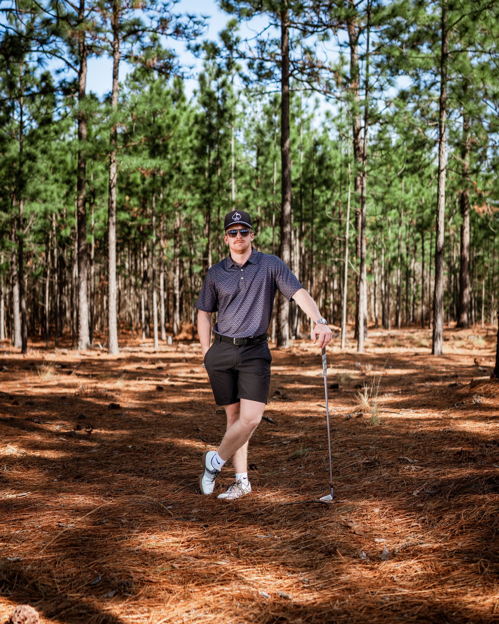 A golfer stands confidently in a forest, wearing a stylish slate mosaic polo and shorts, holding a golf club.
