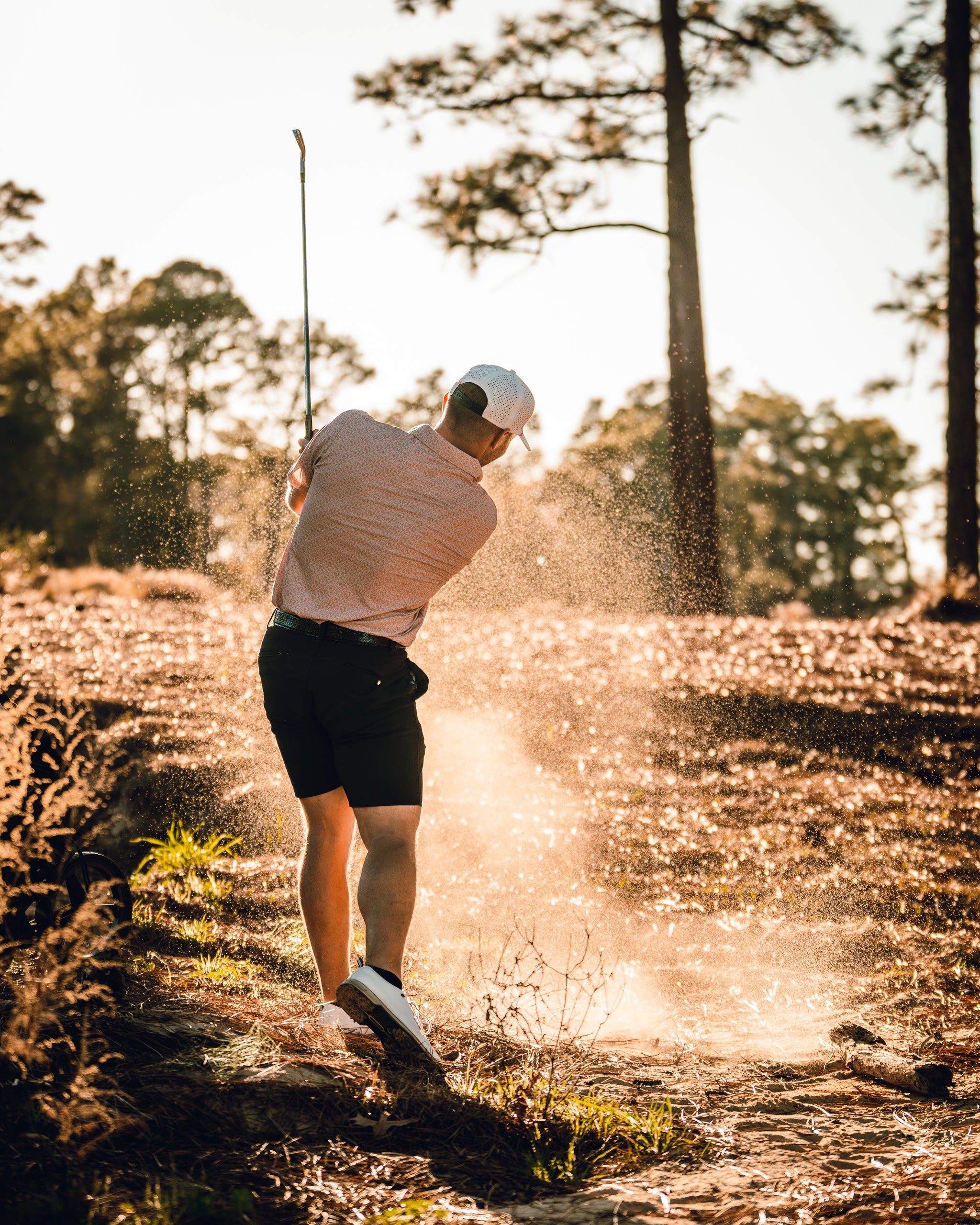 Golfer in a stylish outfit swings a club, sending sand flying from a bunker on a sunlit course surrounded by trees.