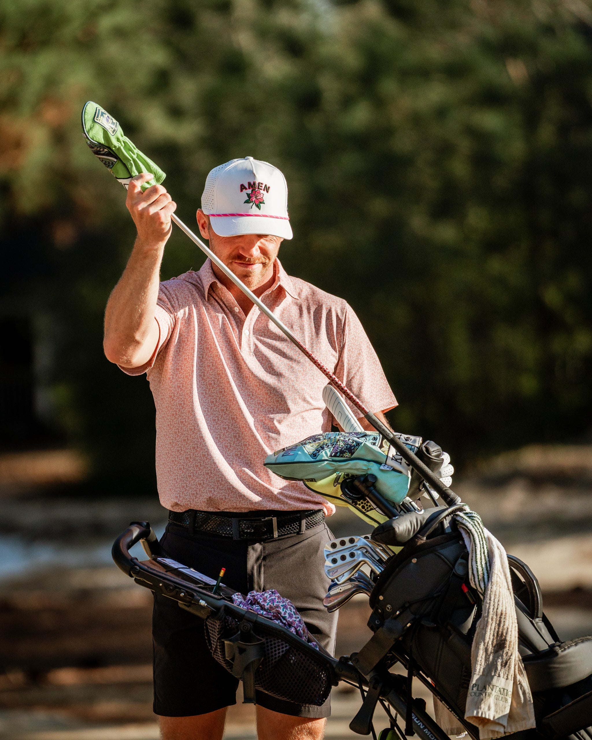 A golfer in a pink patterned shirt and cap inspects a green headcover while standing by his golf bag on the course.